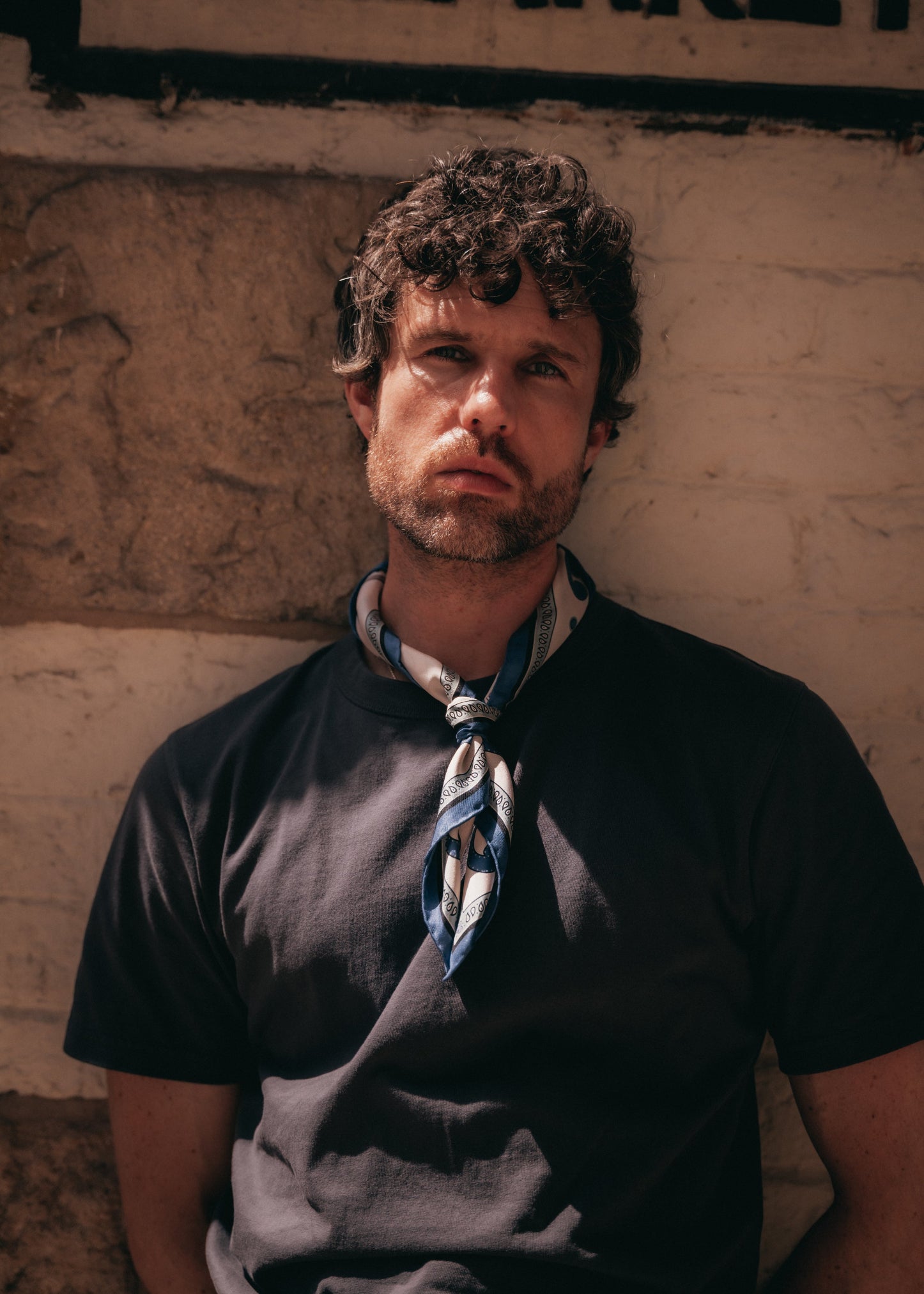 A man wearing a black t-shirt and a 'Belgravia in Cream and Blue' silk neckerchief from SOHO Scarves, leaning against a textured beige wall.