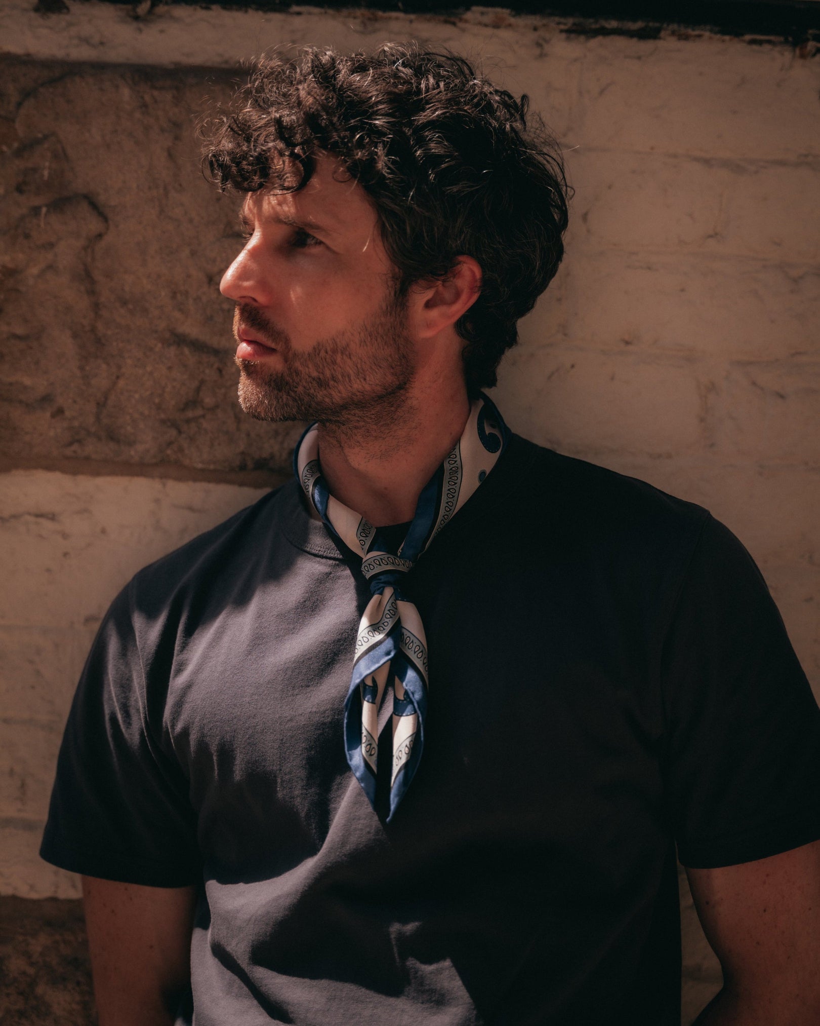 A man looking to his right wearing a black t-shirt and a 'Belgravia in Cream and Blue' silk neckerchief from SOHO Scarves, leaning against a textured beige wall.