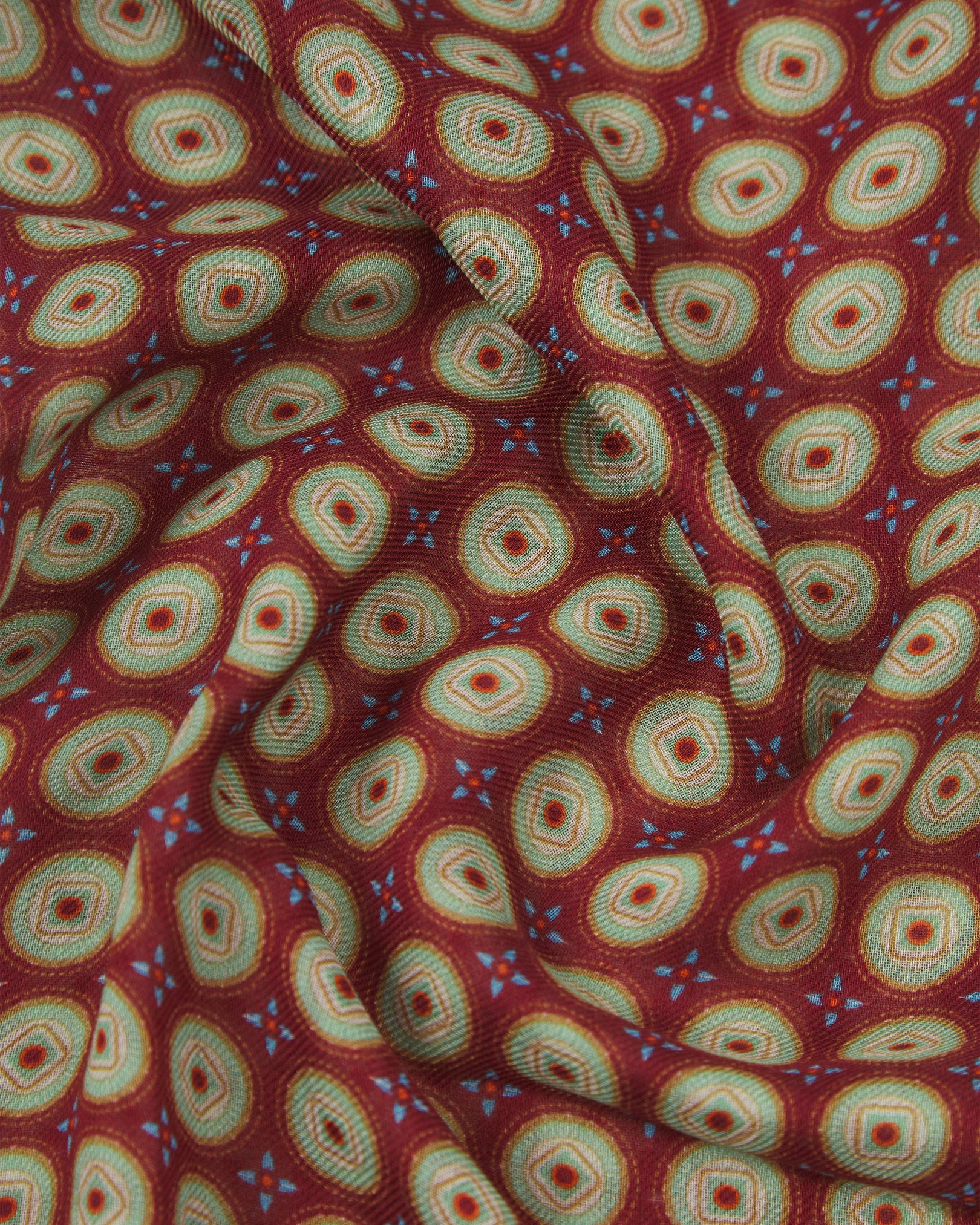A ruffled close-up of the Bellingham 'Gardenia' bohemian wide scarf, showing the soft modal and wool blend fabric with intricate concentric circular motifs in green, gold, orange and blue on a rich maroon background.