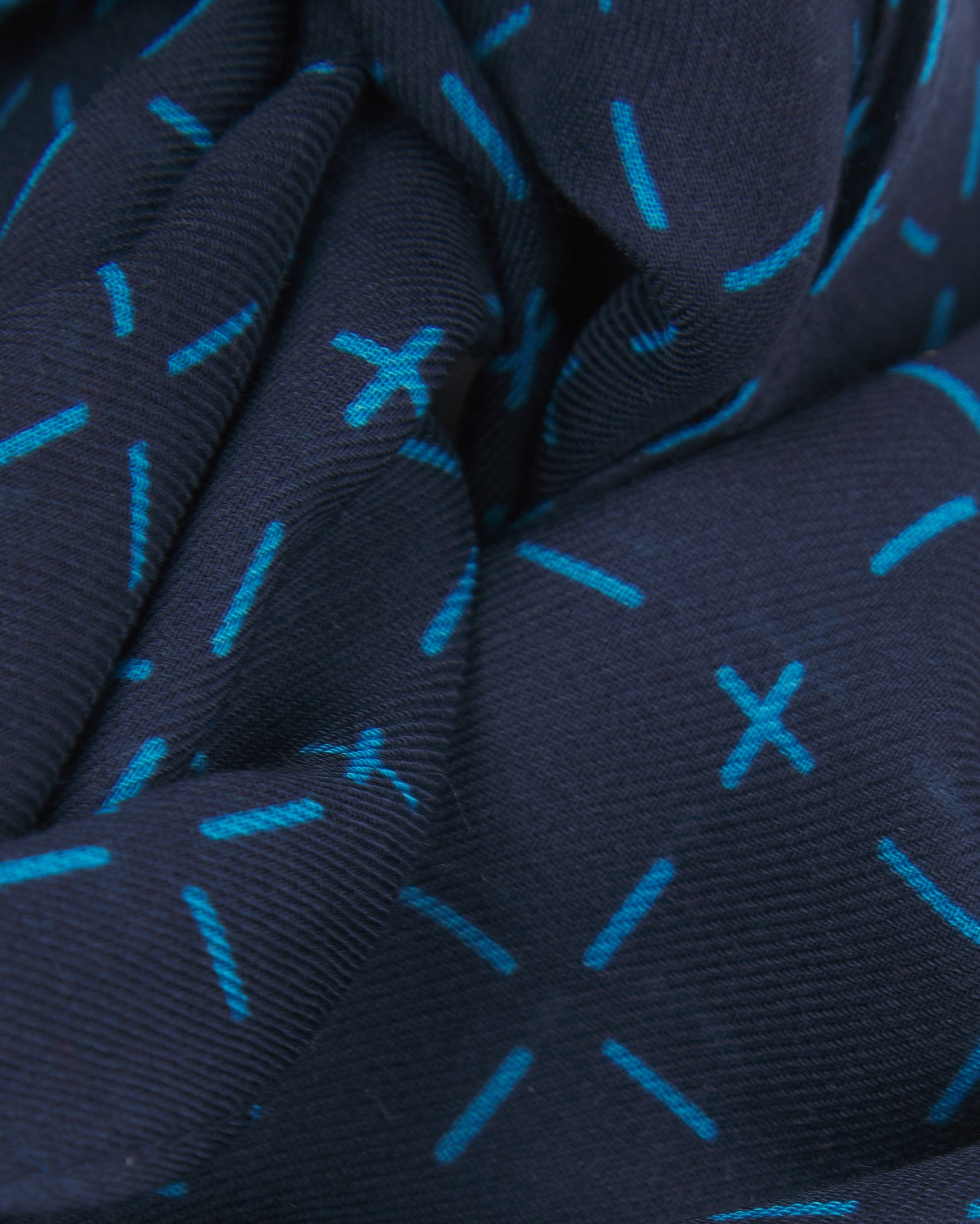A ruffled close-up of the 'Crosses' blue bohemian wide scarf, showing the soft modal and wool blend fabric with a repeating geometric grid of light blue dashes and crosses on a navy blue background.
