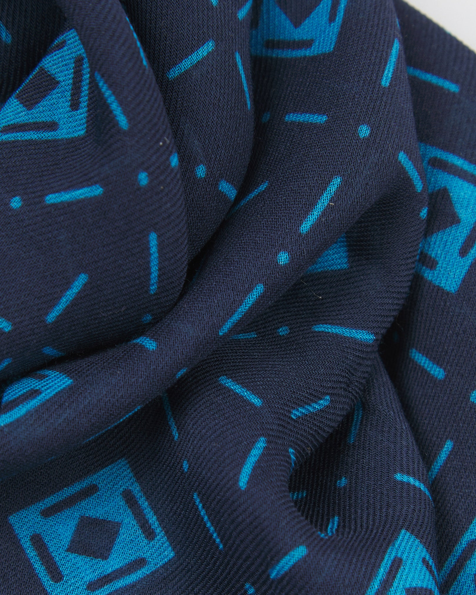 A ruffled close-up of the 'Diamonds' blue bohemian wide scarf, showing the soft modal and wool blend fabric with an ordered diamond mosaic of light blue lozenges and crosshair motifs on a navy blue background.