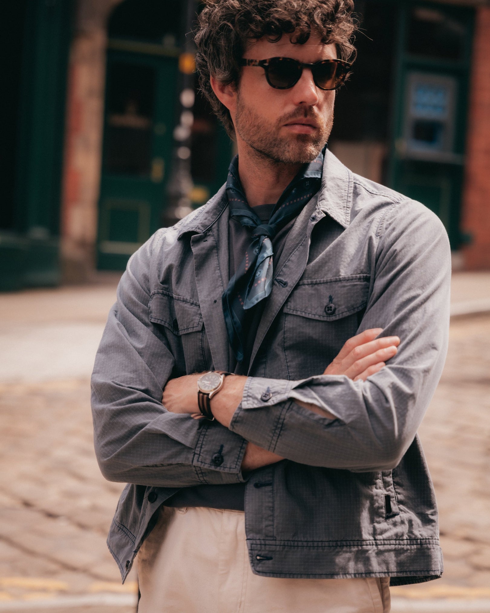 A man wearing a gray denim jacket over a dark shirt and scarf, paired with beige trousers, finished with a loosely tied 'Chelsea' indigo neckerchief from SOHO Scarves.