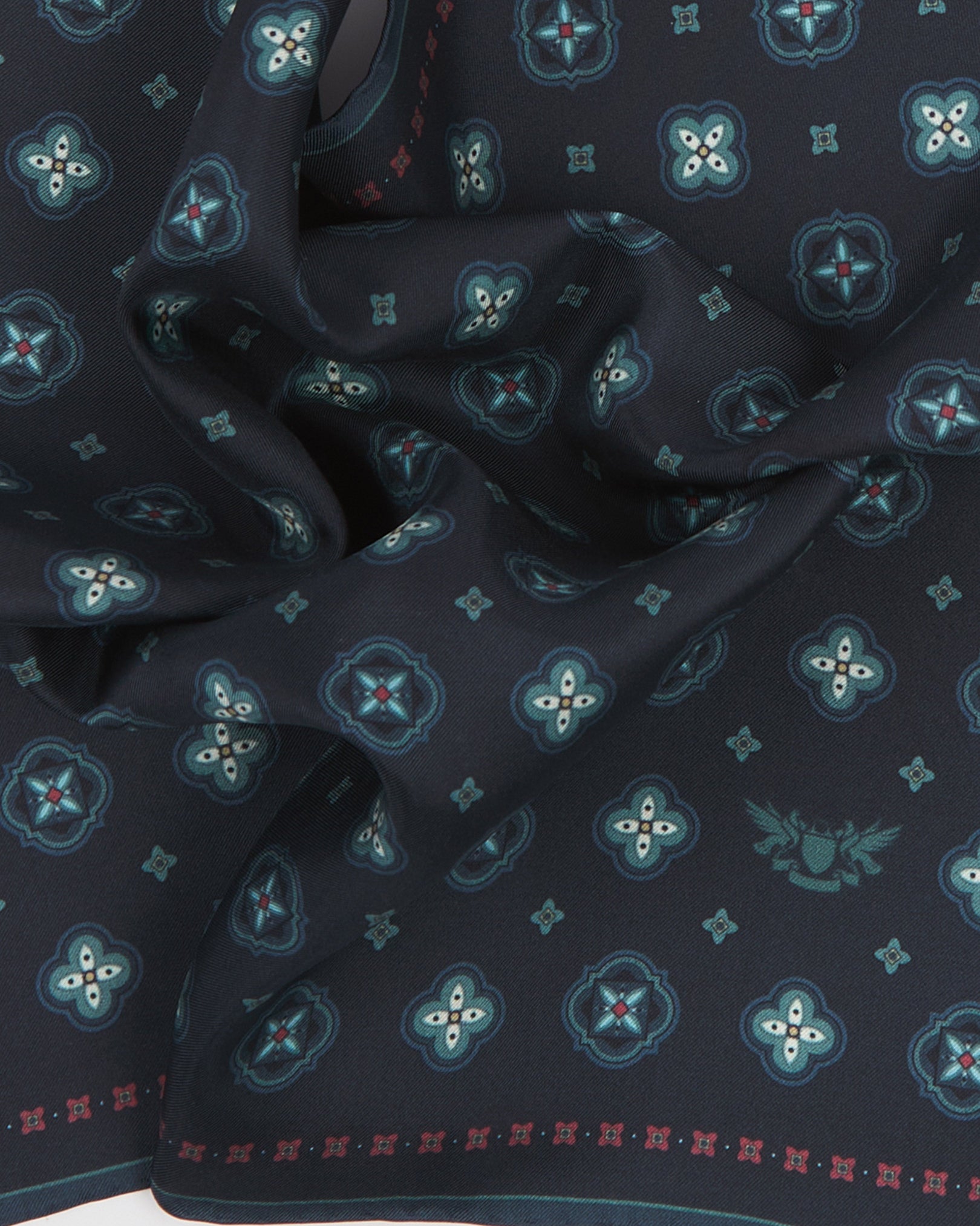 A ruffled close-up of the 'Chelsea' silk pocket square from SOHO Scarves, showing overlapping teal quatrefoil medallions with crimson star accents and pale teal cross motifs on a deep midnight navy ground.