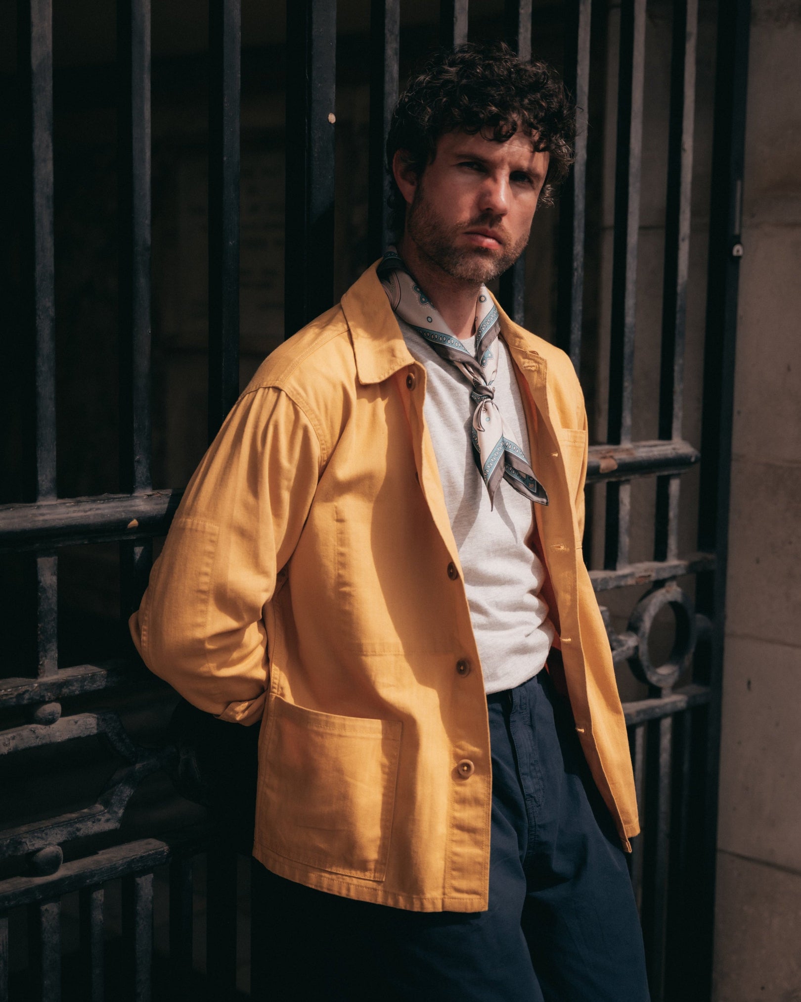 A man wearing a mustard yellow button-up overshirt over a white shirt and a 'Belgravia' pale khaki patterned silk necherchief from SOHO Scarves.