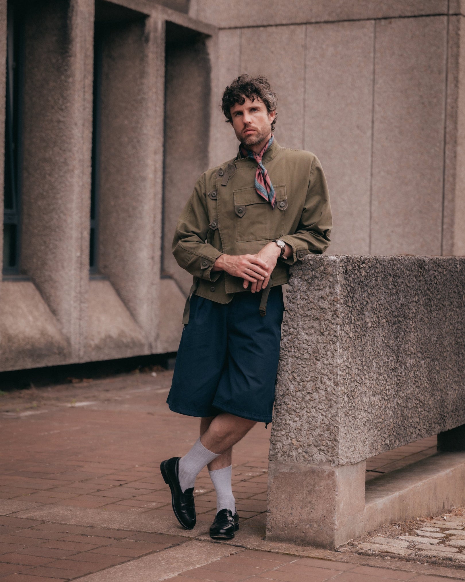 A man wearing a vintage-style olive green jacket, navy shorts, white socks, and black loafers leans against a concrete wall in an urban setting. Tied tightly round the neck is the Chiswick silk neckerchief.