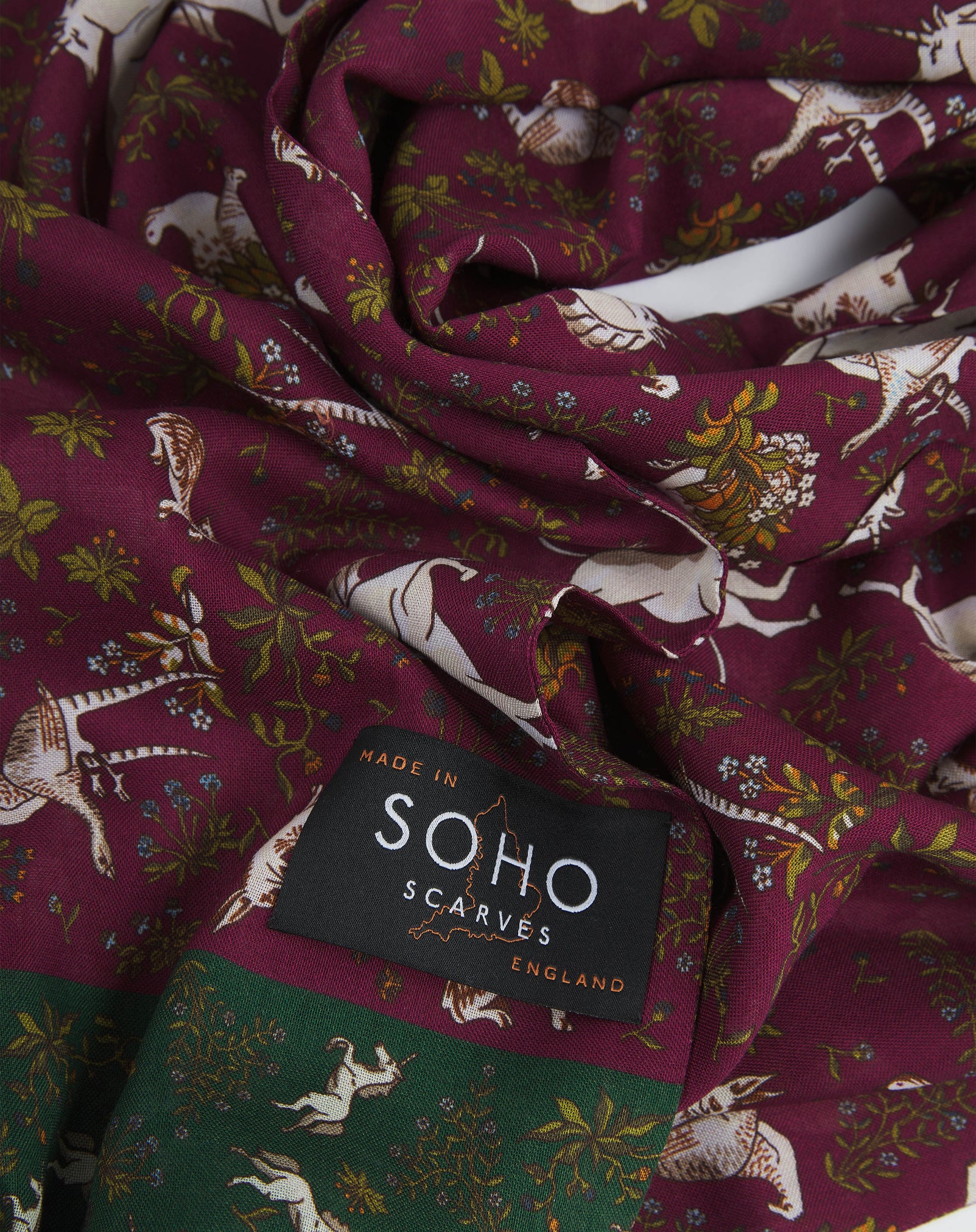 A ruffled, close-up view of the UK-made wine and green silk and wool scarf depicting unicorns, hares and pheasants amid botanical forms. Clearly showing the premium silk and wool weave.