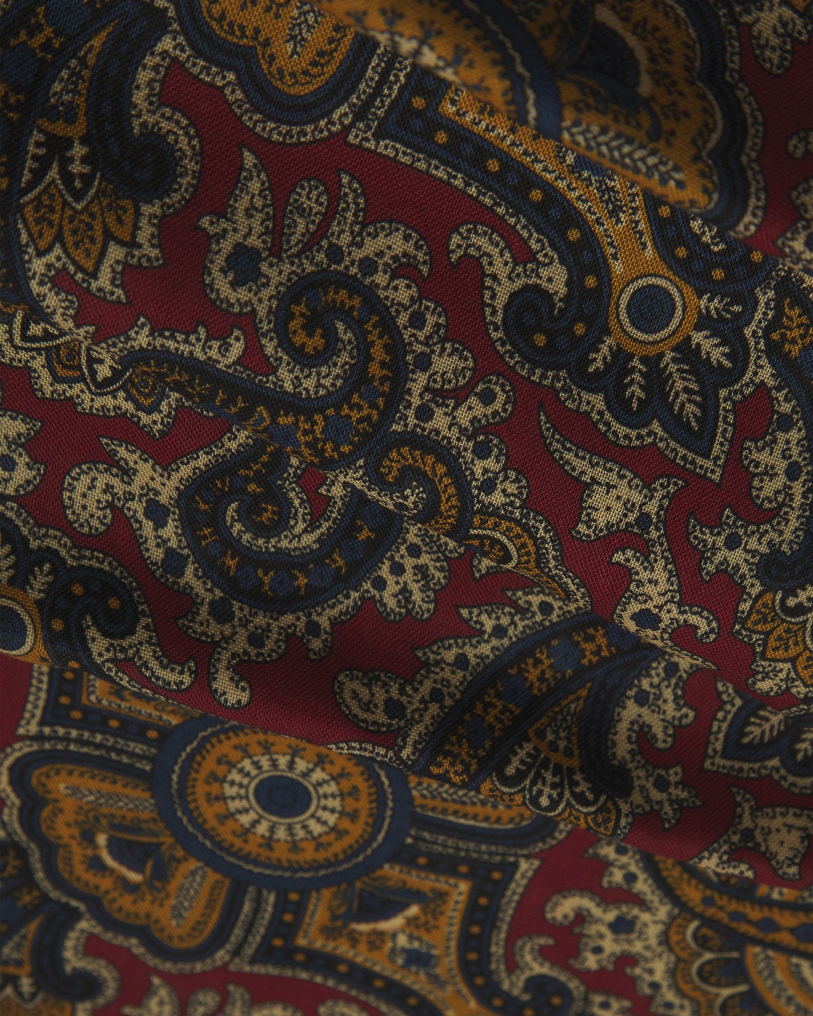 A flat and ruffled close-up view of the UK-made deep red paisley scarf. Clearly showing the premium silk and wool weave and the detailed red, blue and yellow paisley patterns against a red background.