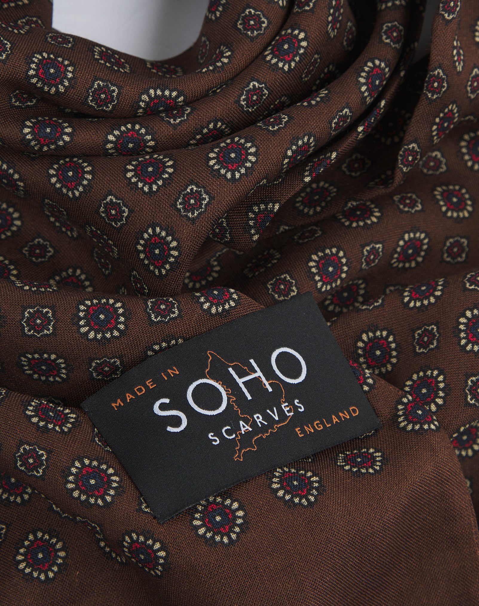 A ruffled, close-up view of the UK-made chocolate-brown silk and wool scarf adorned with floral-inspired repeat patterns in cream and red. Clearly showing the premium silk and wool weave.