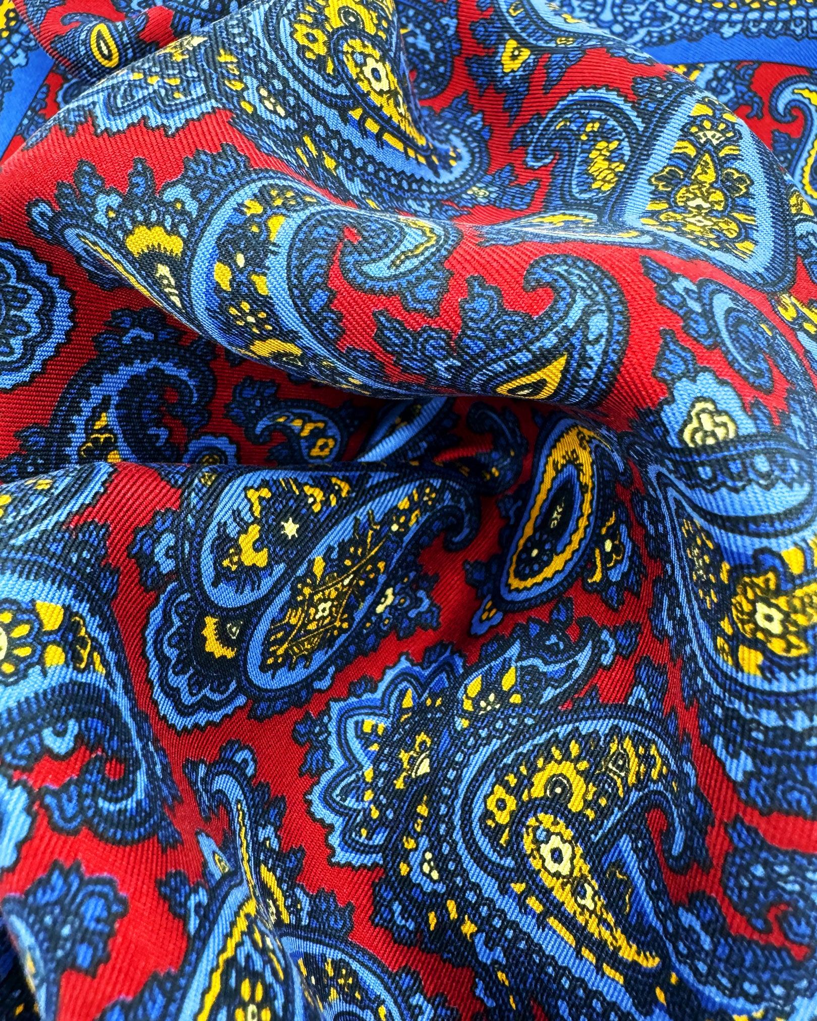 A ruffled close-up of the 'Avebury' pocket square, presenting a closer view of the yellow and blue paisley patterns against the attractive lustre of the madder silk material. 