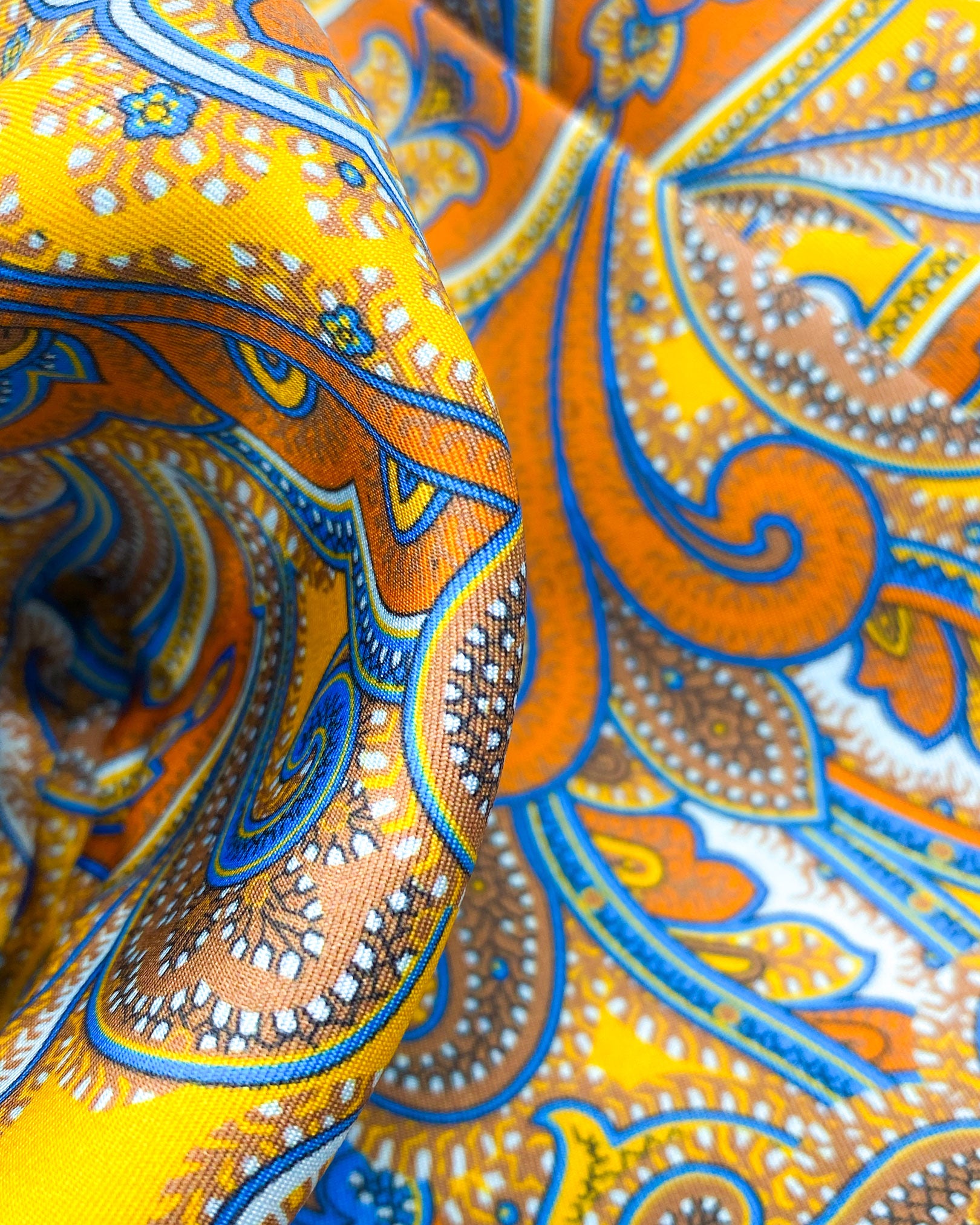 Ruffled close-up view of the 'Carnaby' silk aviator scarf, presenting a closer view of the swirls of warm paisley tones against a bright golden ground and lustre of the silk material.
