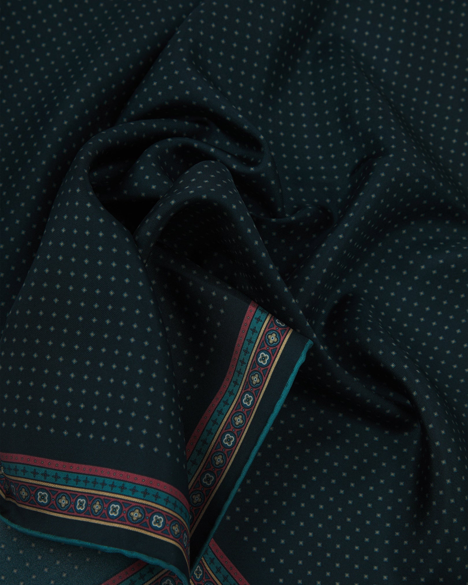 A ruffled close-up of the 'Chiswick' silk neckerchief from SOHO Scarves, featuring a fine teal micro-dot diamond lattice on a deep midnight indigo ground with a finely woven silk texture and an ornate berry-red and teal geometric medallion border.