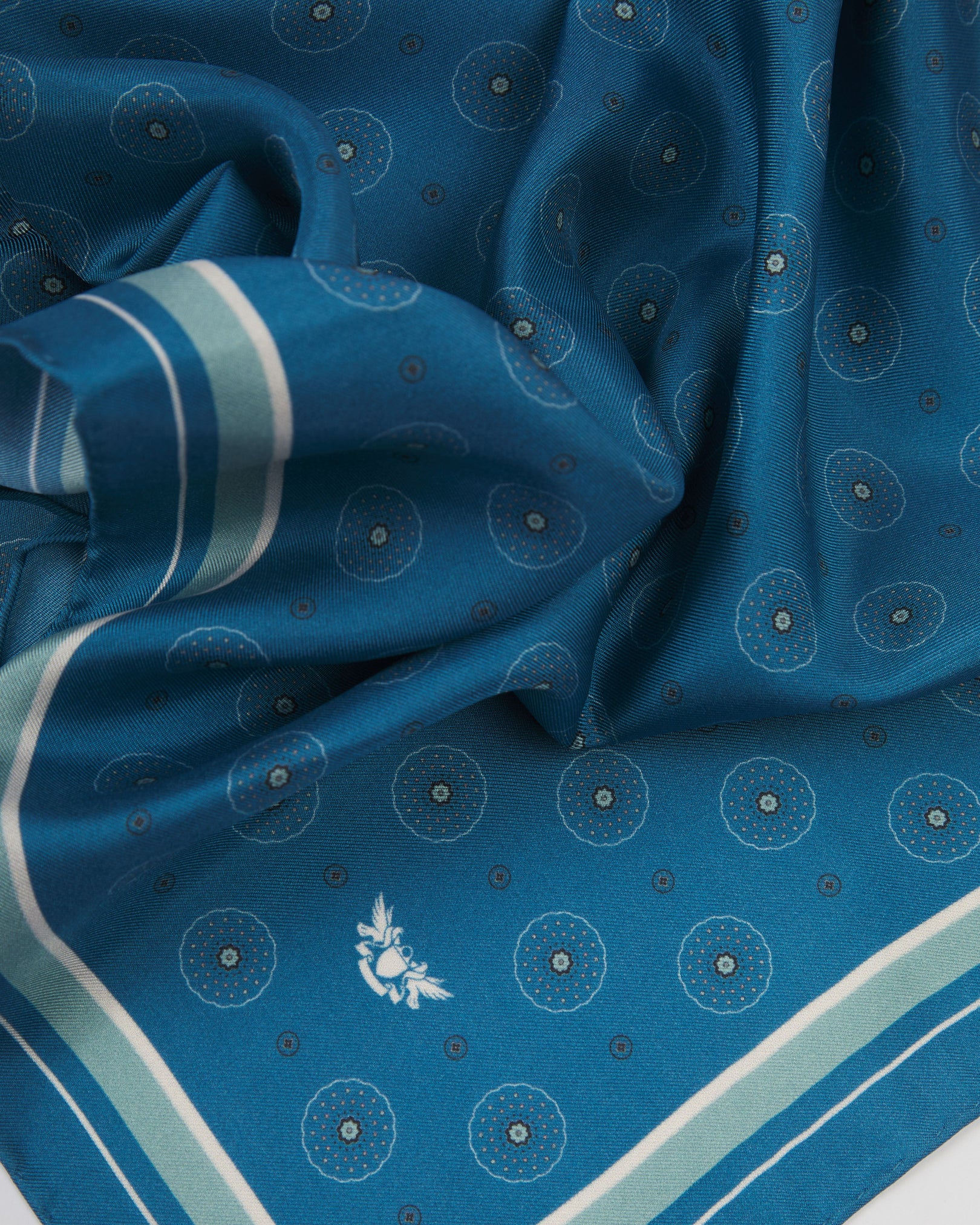 A ruffled close-up of the 'Islington' silk neckerchief from SOHO Scarves, featuring an intricate geometric circle and medallion pattern on a cornflower-slate blue ground, complemented by mint-sage and soft blue-grey borders with a finely woven silk texture.