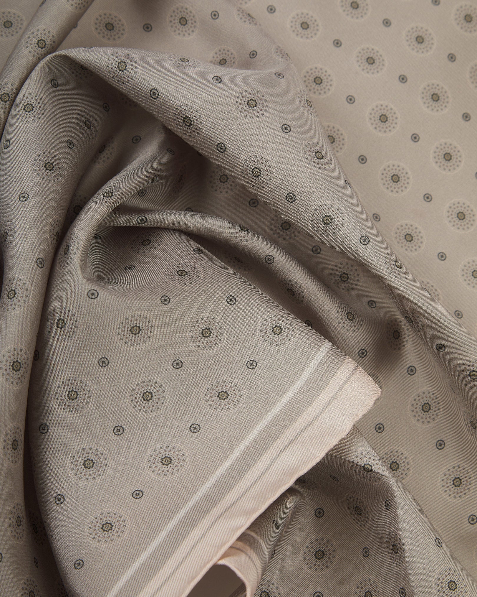 A ruffled close-up of the 'Islington' silk neckerchief from SOHO Scarves, revealing a tonal geometric circle and sunburst pattern on a warm greige-stone ground with a finely woven silk texture and elegant cream border detail.