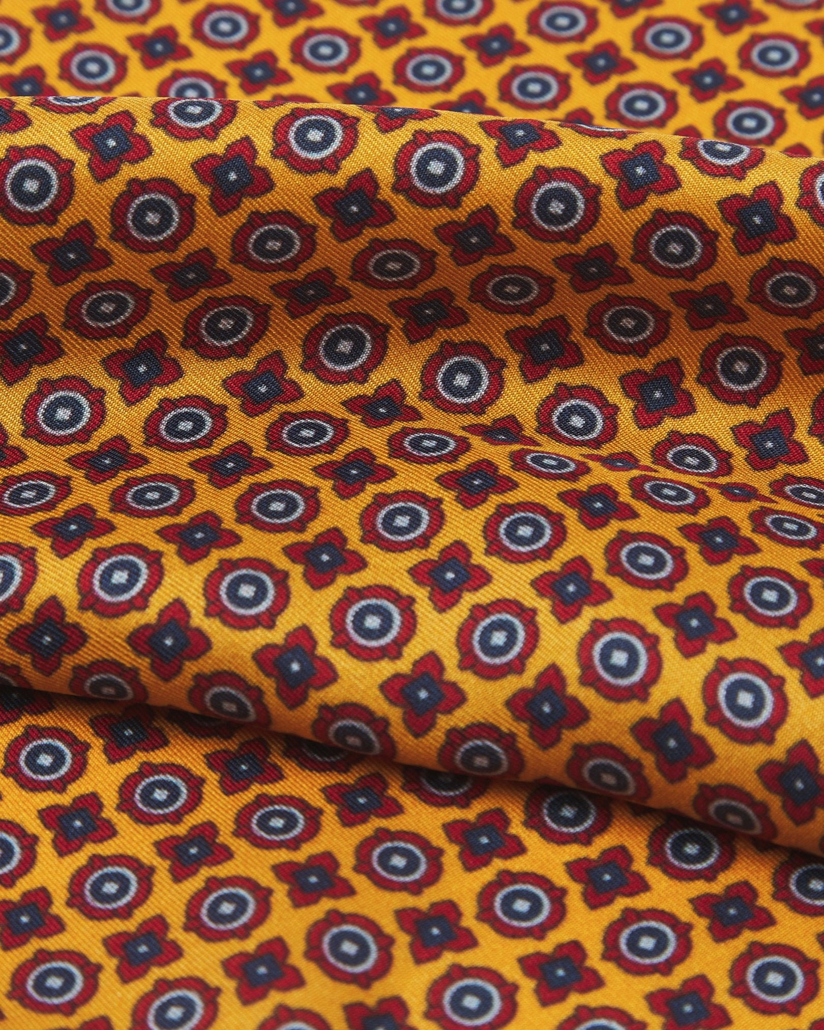 A ruffled close-up of the 'Toshima' silk neckerchief from SOHO Scarves, presenting a closer view of the small blue and red stylised floral patterns on a golden-orange ground.