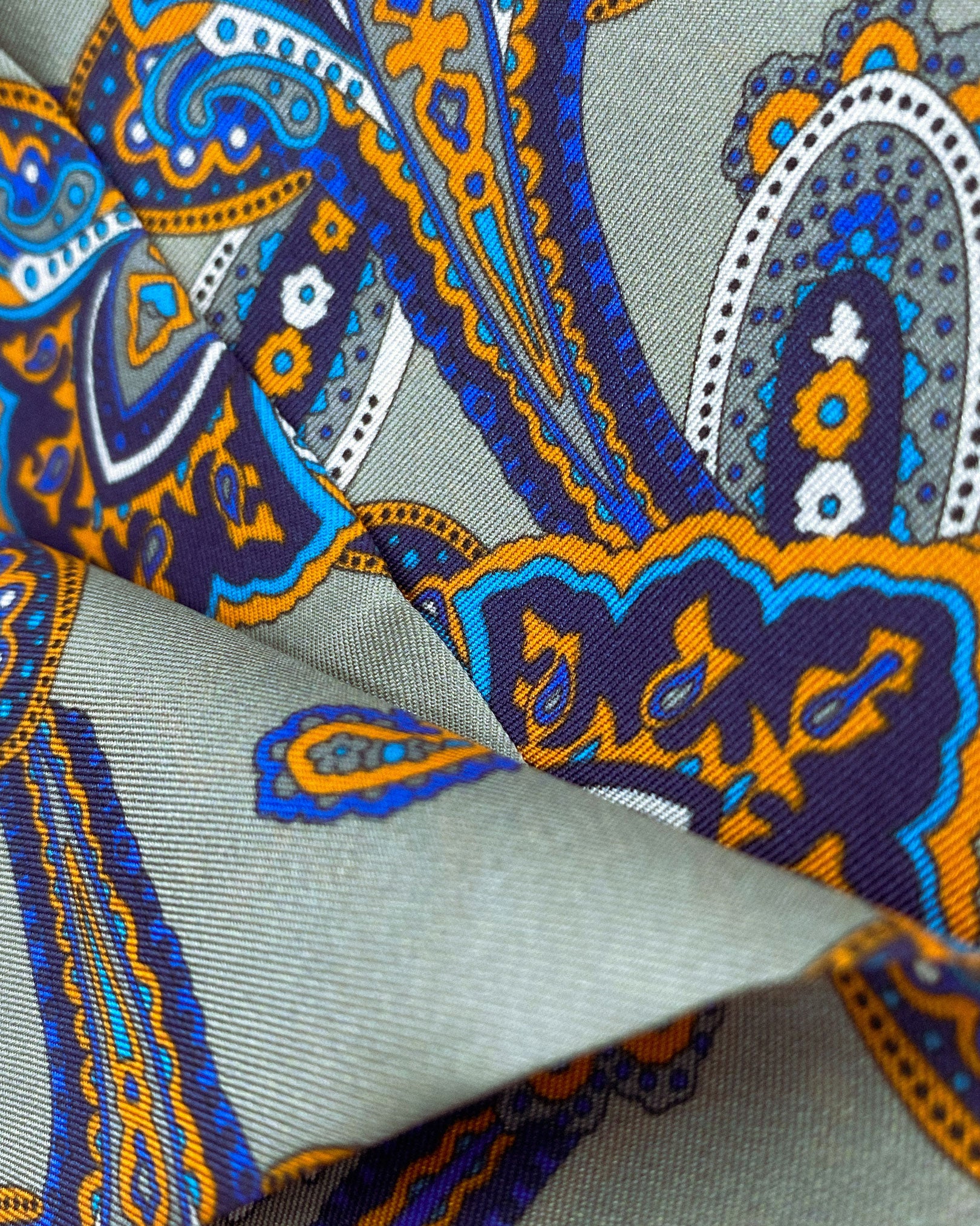 Ruffled close-up view of the Tremblant polyester scarf, presenting a closer view of the  orange and light blue paisley forms on a grey-cream ground.
