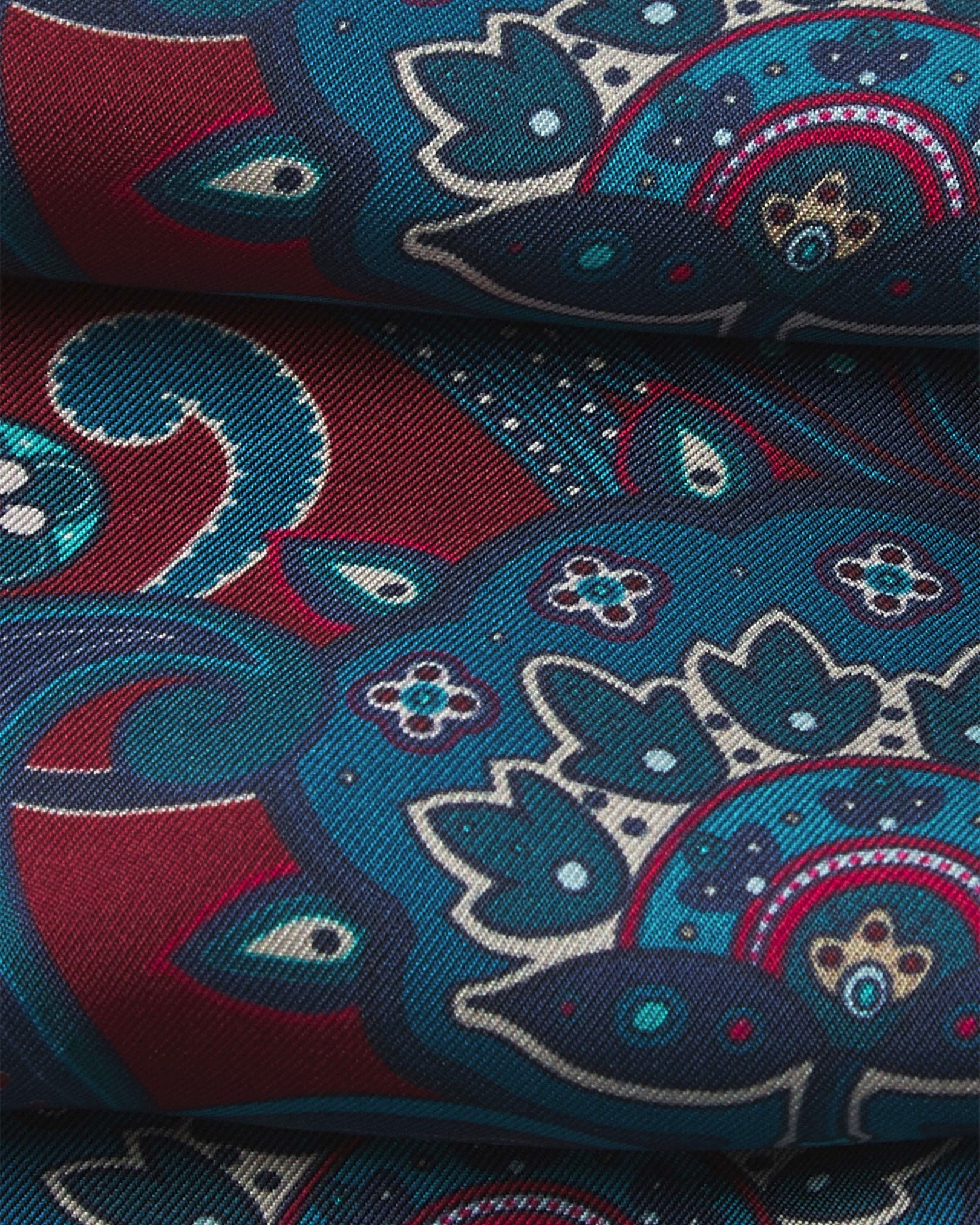 A ruffled close-up of the 'Windermere' silk neckerchief from SOHO Scarves, showing overlapping elements of the blue and blue-green paisley patterns against a rust-red background.