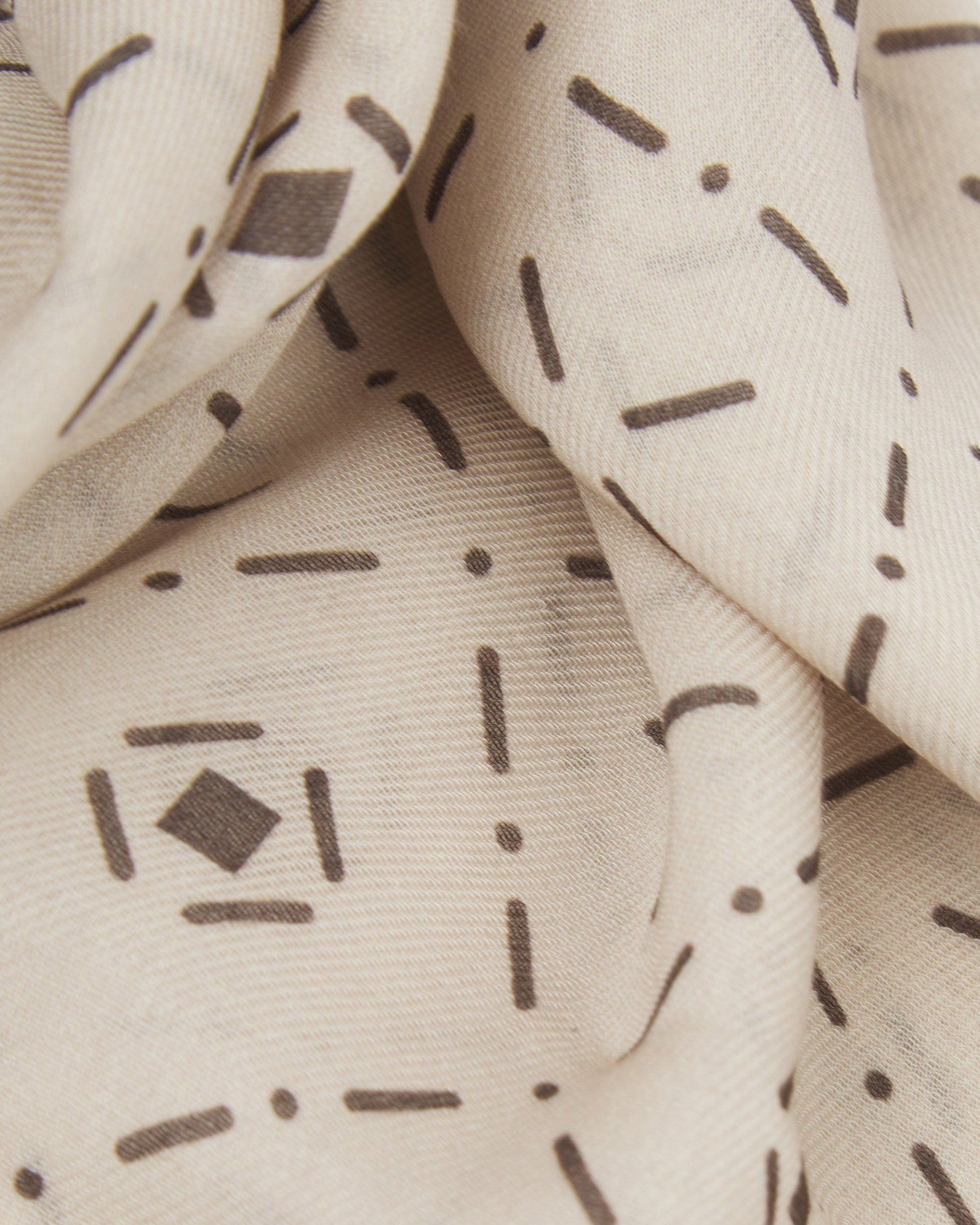 A ruffled close-up of the 'Diamonds' cream bohemian wide scarf, showing the soft modal and wool blend fabric with an ordered diamond mosaic of dark grey lozenges and crosshair motifs on a pale cream background.