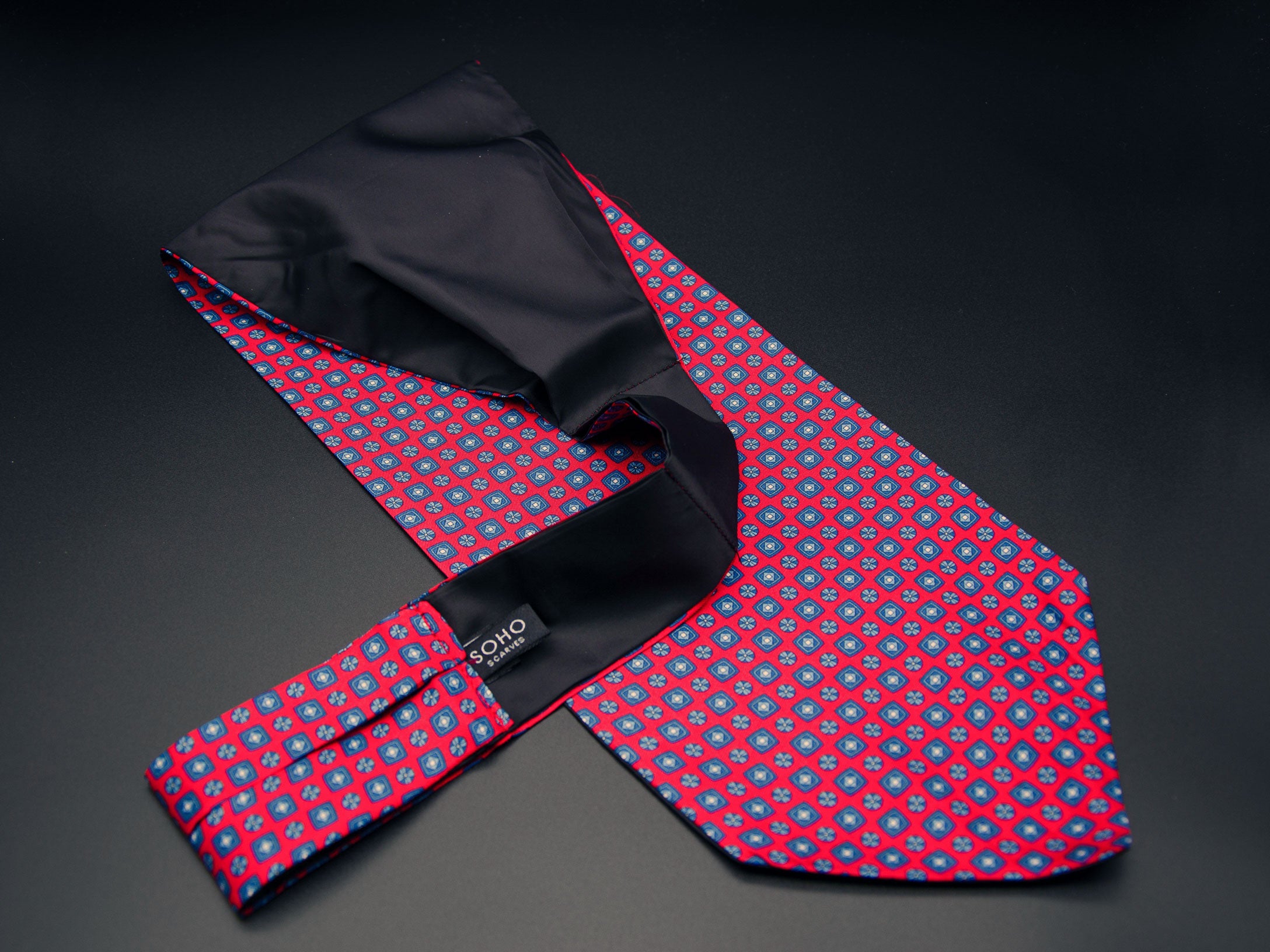 Bright red 'Canaria' single pointed Ascot tie arranged diagonally with wide point in foreground with clear view of blue motifs made up of tiny squares, disks and diamonds. Narrow end of tie folded back and over to reveal dark lining and 'SOHO Scarves' branding label.