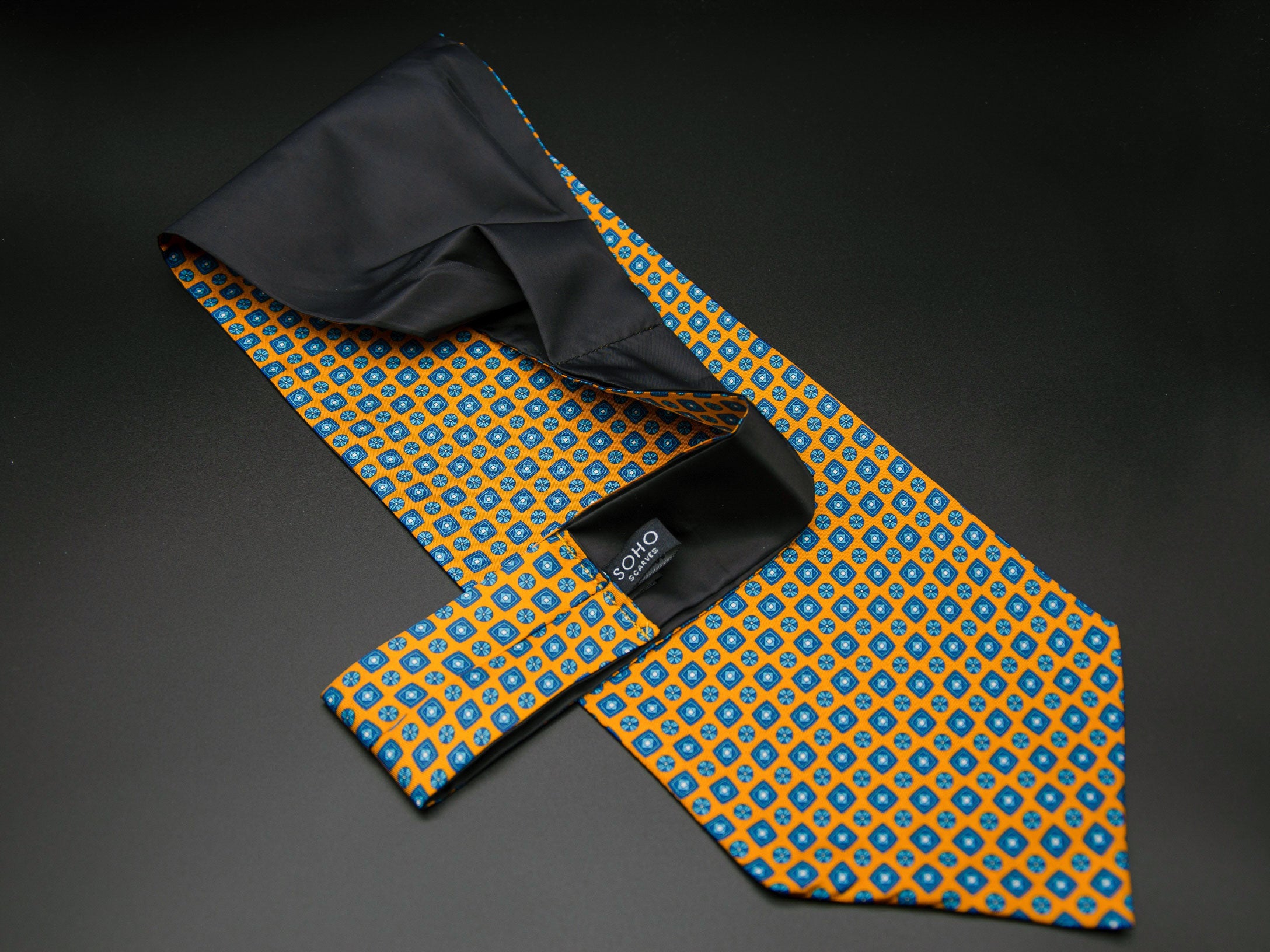 Golden 'Madrid' single pointed Ascot tie arranged diagonally with wide point in foreground with clear view of blue motifs made up of tiny squares, disks and diamonds. Narrow end of tie folded back and over to reveal dark lining and 'SOHO Scarves' branding label.