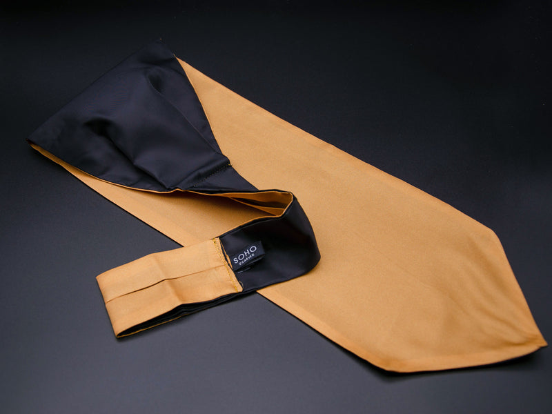 Golden 'Mijas' single pointed Ascot tie arranged diagonally with wide point in foreground. Narrow end of tie folded back and over to reveal dark lining and 'SOHO Scarves' branding label.
