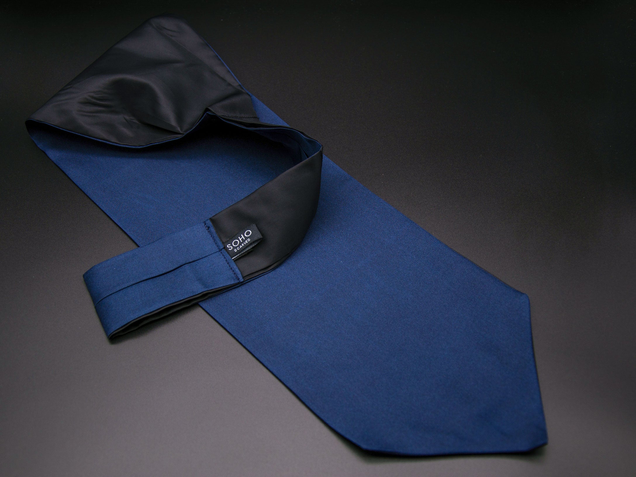 Blue 'Sebastian' single pointed Ascot tie arranged diagonally with wide point in foreground. Narrow end of tie folded back and over to reveal dark lining and 'SOHO Scarves' branding label.
