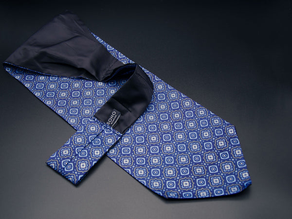 Blue 'Seville' single pointed Ascot tie arranged diagonally with wide point in foreground and clear view of pale blue motifs made up of offset floral-inspired diamond patterns with golden accents. Narrow end of tie folded back and over to reveal dark lining and 'SOHO Scarves' branding label.
