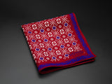 Wool 'Hampi' pocket square folded showing white and blue decorative floral patterns aranged in linear repeats on a red ground. Framed by an intricate deep blue border. SOHO Scarves branding clearly visible on bottom-right.