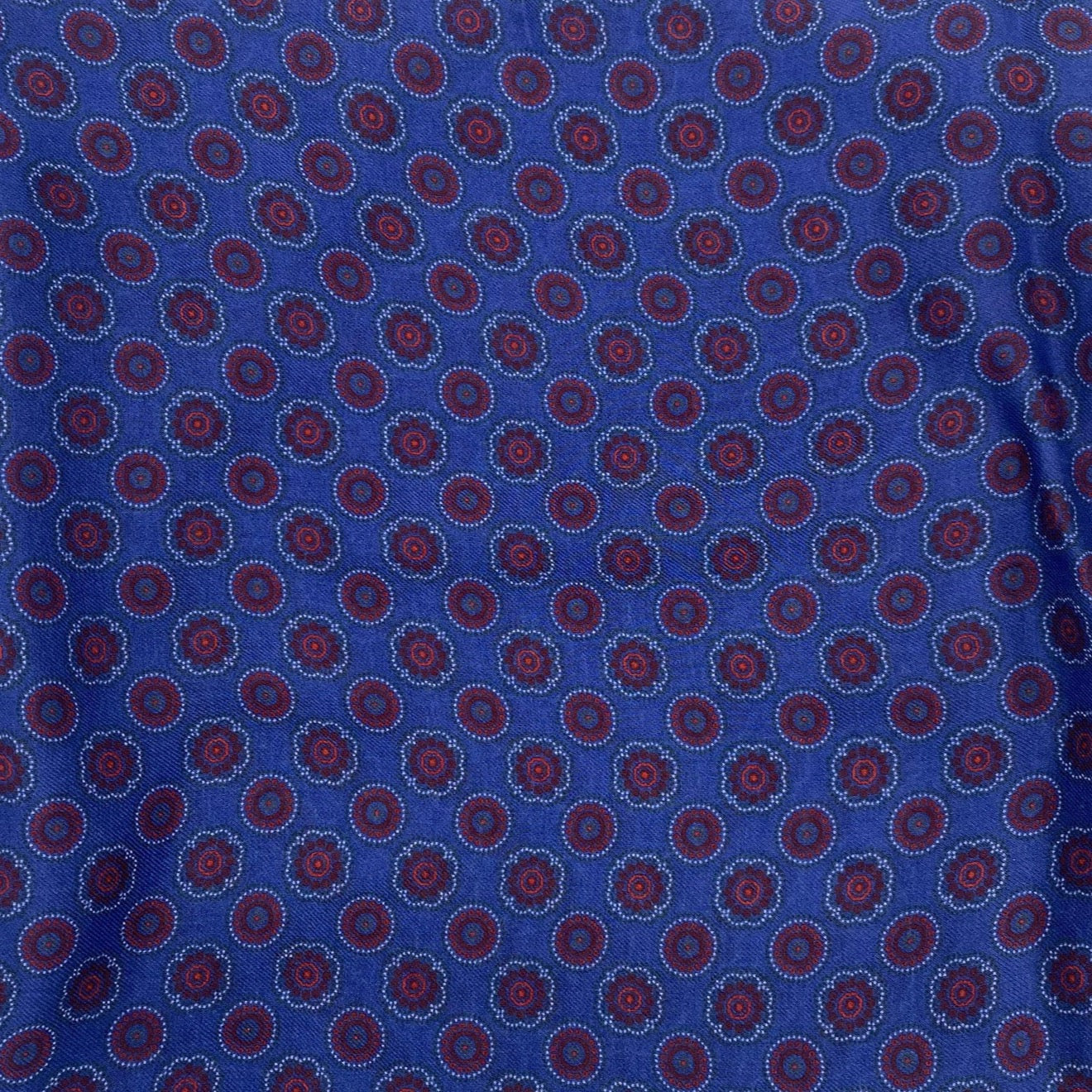 A flat, folded view of 'The Bellevue' boho wide scarf on a blue background. Clearly showing the circular red patterns and floral inspired patterns.