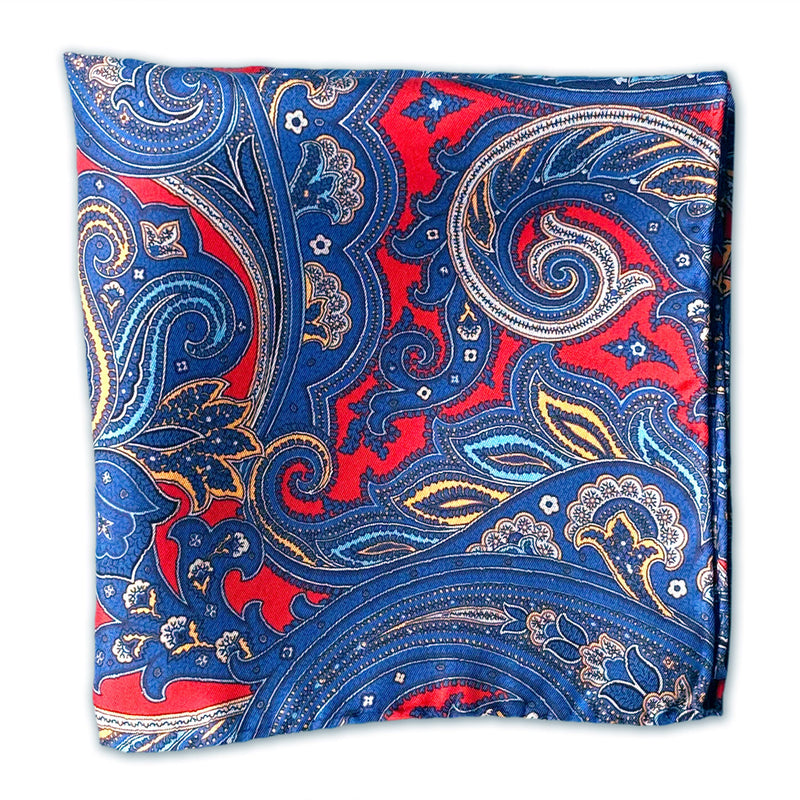 Flat view of the 'Oxford' large paisley patterned pocket square, folded over into a smaller sqaure shape. Showing the large and smaller paisley pattern and surrounding floral patterns on a deep red ground.