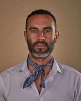 Portrait view of model looking straight ahead, wearing 'The DeZon' blue patterned neckerchief on a navy ground. Tied in a simple front-facing knot.