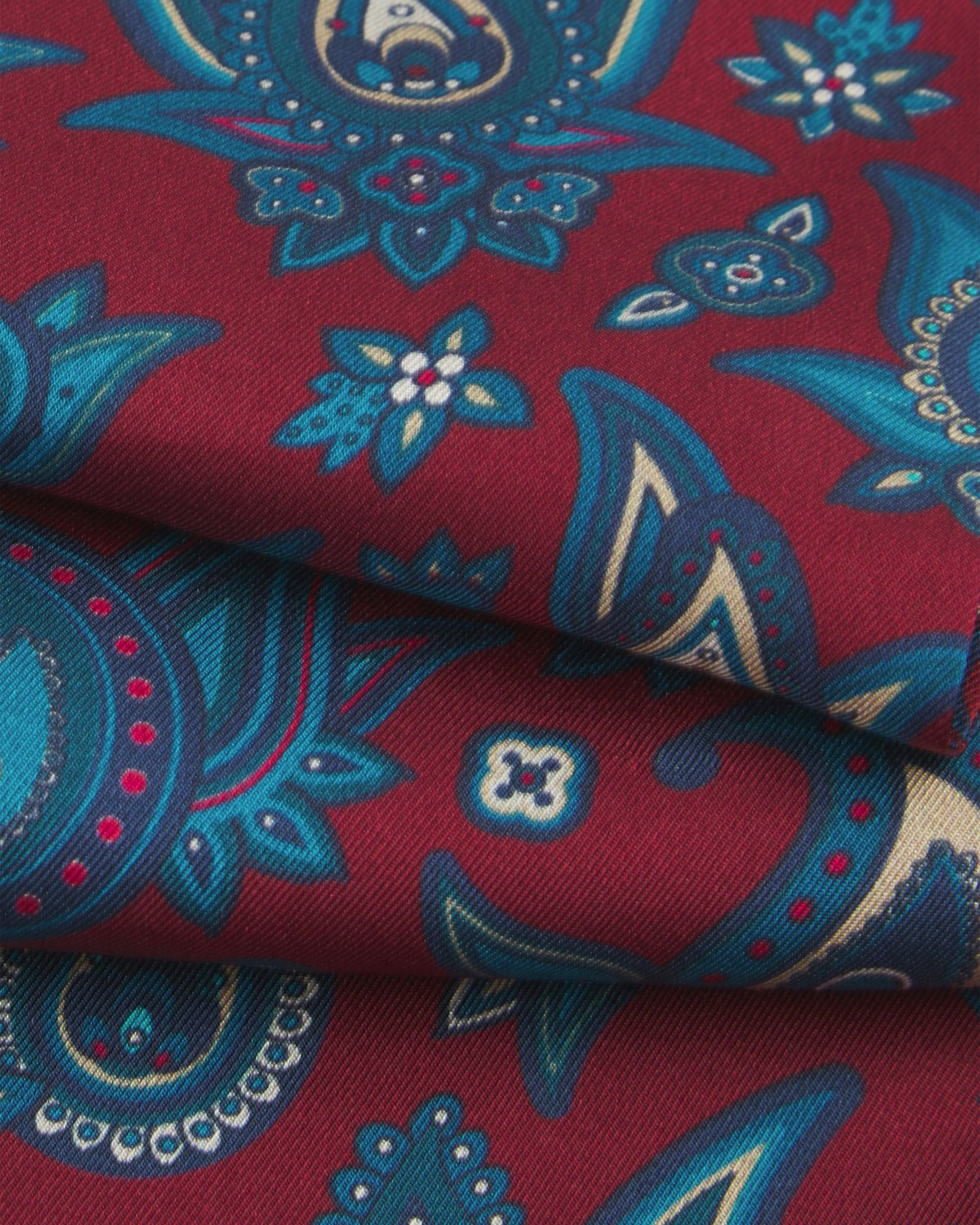 A ruffled close-up of the 'Grassmere' rust silk scarf, presenting the overlapping elements of blue and blue-green paisley patterns.