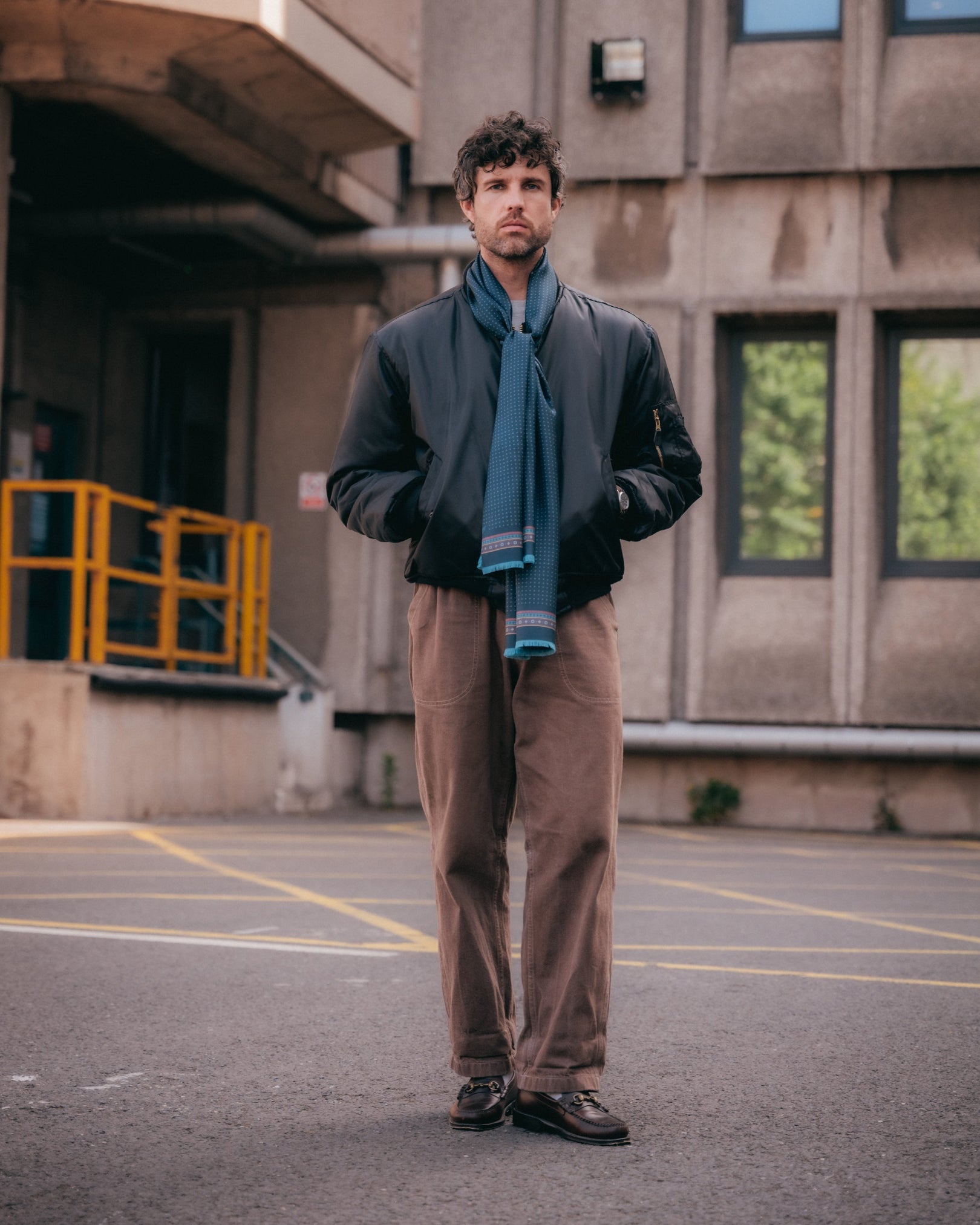 Full-length view of man outside a brutalist concrete building wearing the indigo 'Chiswick' silk scarf from SOHO Scarves. Tied loosely around the neck and paired with brown jeans and a navy bomber jacket.
