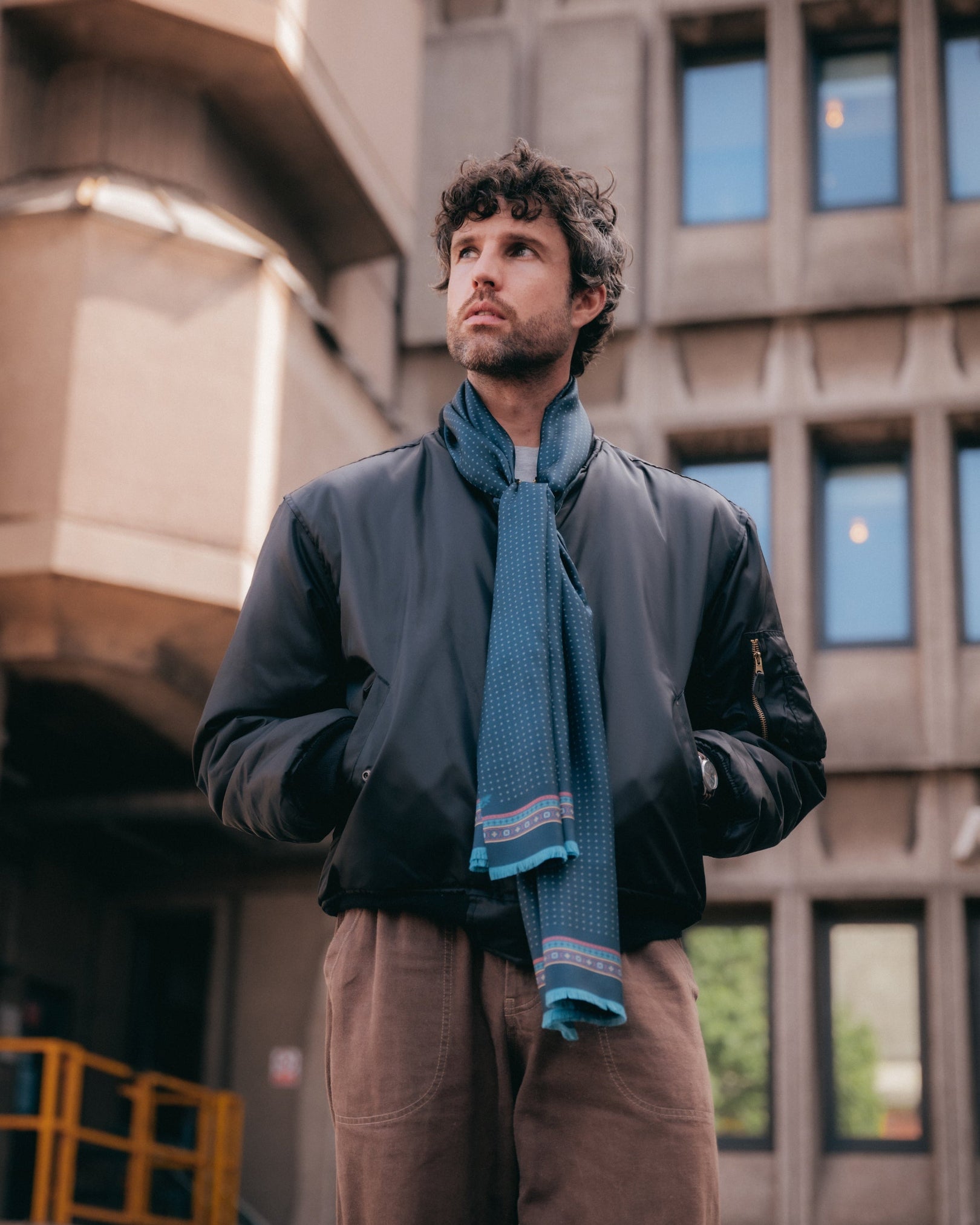 Top-half view of man wearing the indigo 'Chiswick' silk scarf loosely around the neck. Paired with brown jeans and a navy bomber jacket.