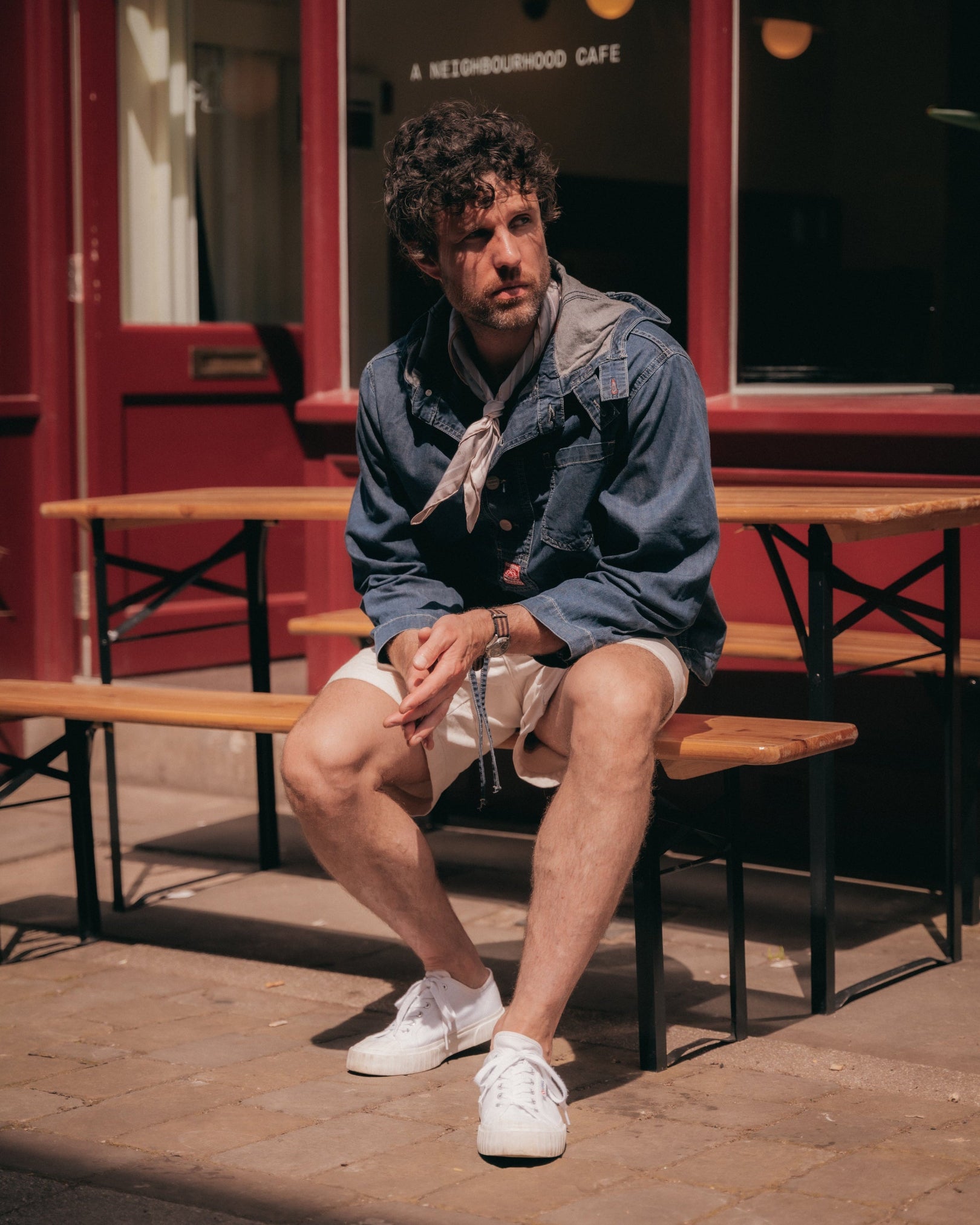 Man seated outside a café wearing the 'Islington' warm grey silk neckerchief loosely around the neck, paired with light beige shorts and a denim jacket