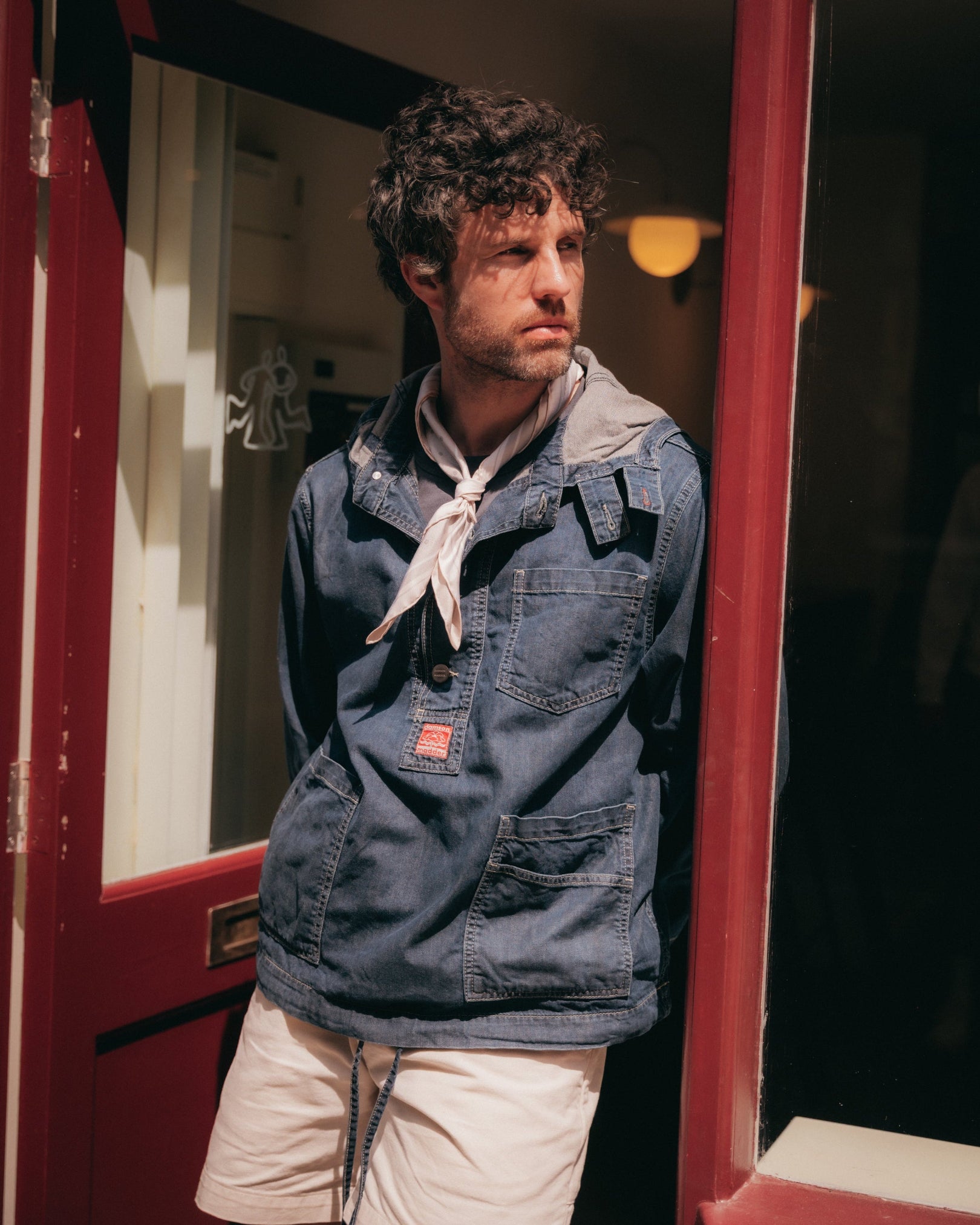 A man wearing a blue denim utility jacket with the 'Islington' warm grey silk neckerchief and light beige shorts, standing in a doorway with red trim.