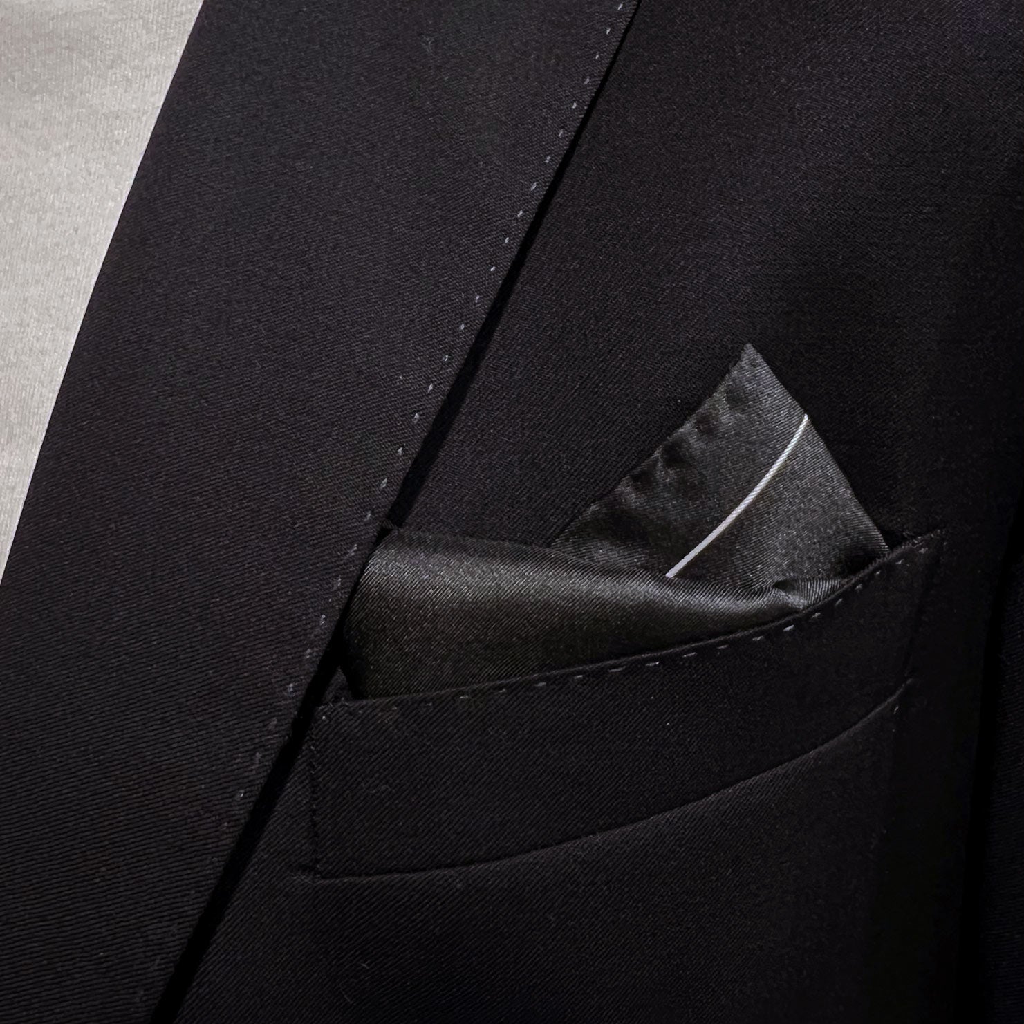 Close-up of black 'Chaplin' silk pocket square in breast pocket of dark suit jacket.