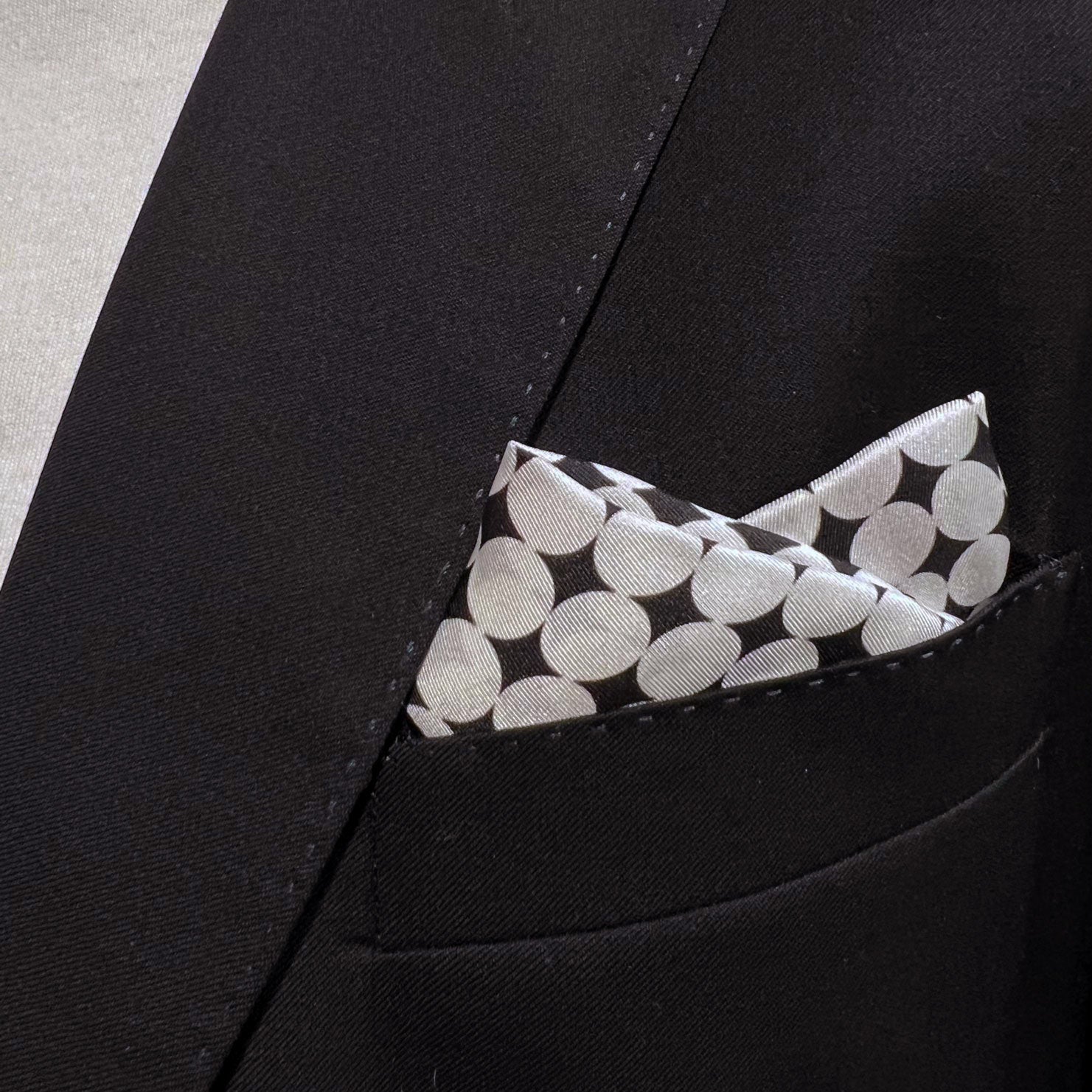 Close-up of  'Keaton' silk pocket square with white disc patterns in breast pocket of dark suit jacket.