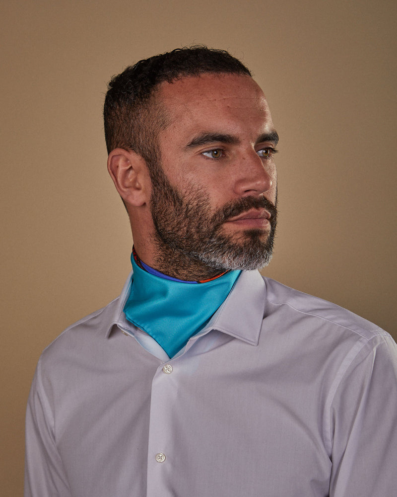 Head and shoulders view of model facing his left, wearing the 'BT Blue' silk neckerchief with knot tie at the back. Neatly tucked under white shirt.