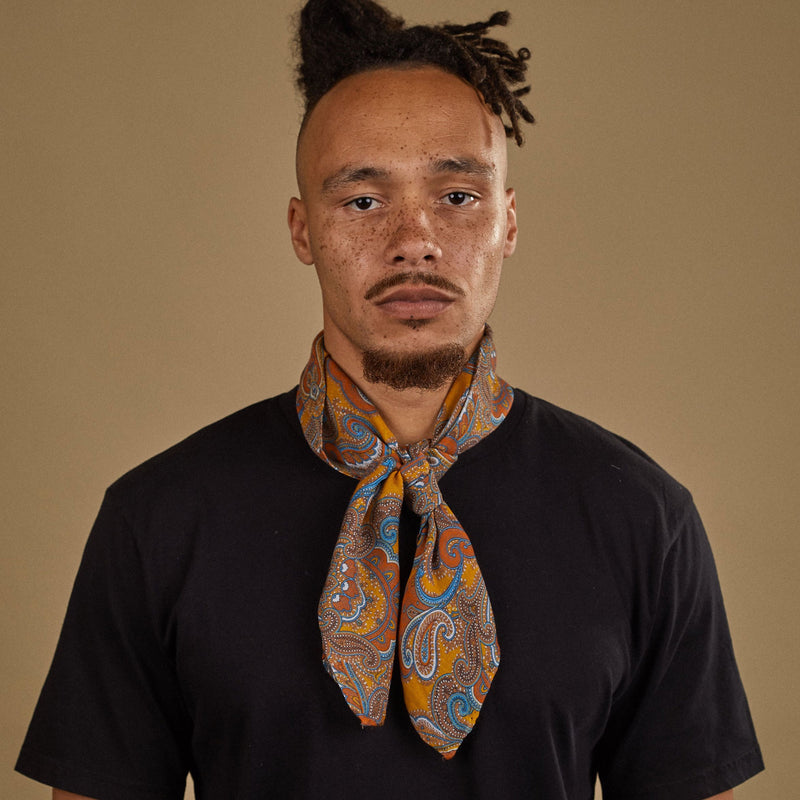 Top-half view of model looking straight ahead wearing 'The Carnaby' multicoloured paisley bandana on a golden ground. Tied snugly around neck in a simple knot.