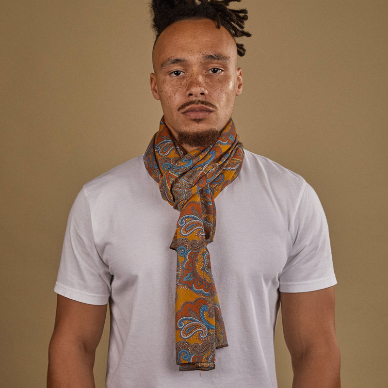 Close-up of model looking straight ahead wearing 'The Carnaby' orange, brown and blue paisley patterned wide bohemian scarf on a gold ground. Wrapped snugly around neck in a simple knot.