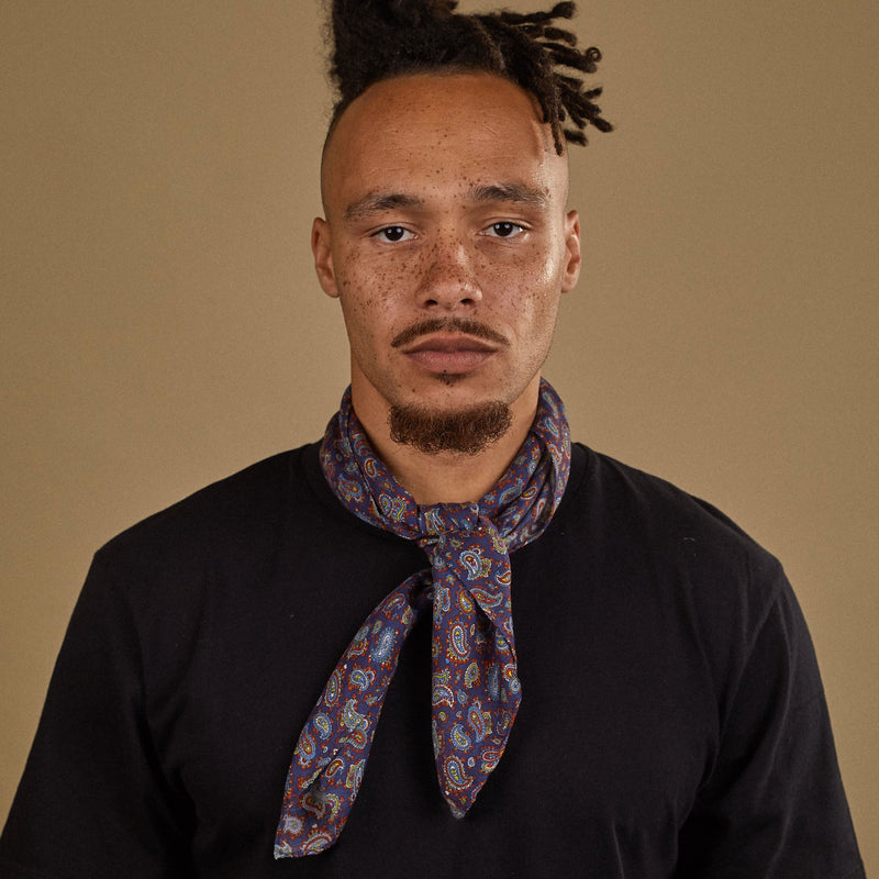 Top-half view of model looking straight ahead wearing 'The Lexington' multicoloured paisley bandana on a deep blue ground. Tied snugly around neck in a simple knot.