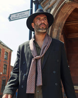 Top-half outdoor shot of model wearing 'The Morne' polyester, mosaic scarf loosely round neck and paired with dark hat and suit jacket.