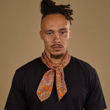Top-half view of model looking straight ahead wearing 'The Niagra' orange and pink floral bandana. Tied snugly around neck in a simple knot and paired with a black t-shirt.