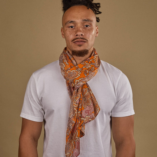 Close-up of model looking straight ahead wearing 'The Niagra' orange and pink floral patterned wide bohemian scarf. Wrapped snugly around neck in a simple knot and paired with a white t-shirt.