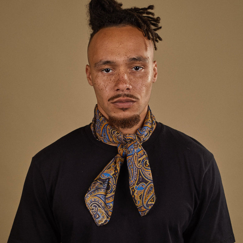 Top-half view of model looking straight ahead wearing 'The Ormond' multicoloured paisley and black bandana. Tied snugly around neck in a simple knot and paired with a black t-shirt.