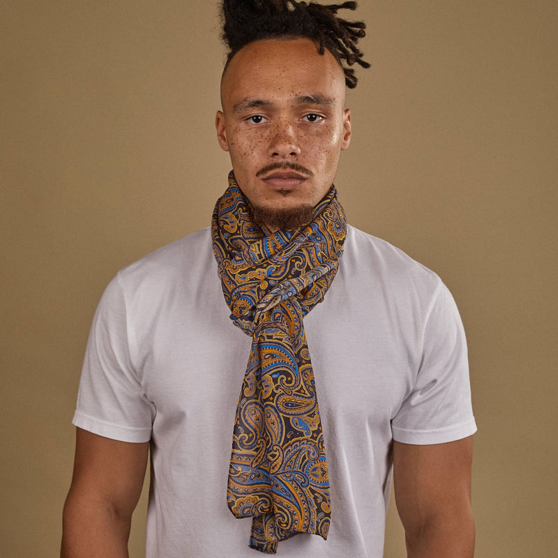 Close-up of model looking straight ahead wearing 'The Ormond' blue, yellow and gold paisley wide bohemian scarf. Wrapped snugly around neck in a simple knot and paired with a white t-shirt.