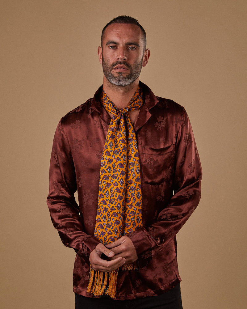 Top-half view of model wearing the orange 'Oviedo' aviator scarf paired with copper-brown silk shirt. Full view of matching orange 3-inch fringe.