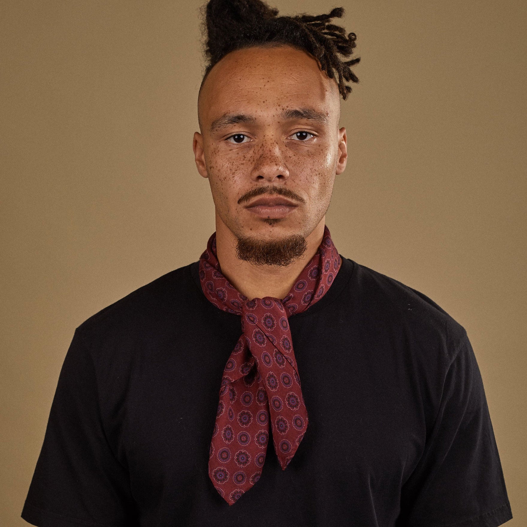 Top-half view of model looking straight ahead wearing 'The Seattle' burgundy and blue geometric bandana. Tied snugly around neck in a simple knot and paired with a black t-shirt.