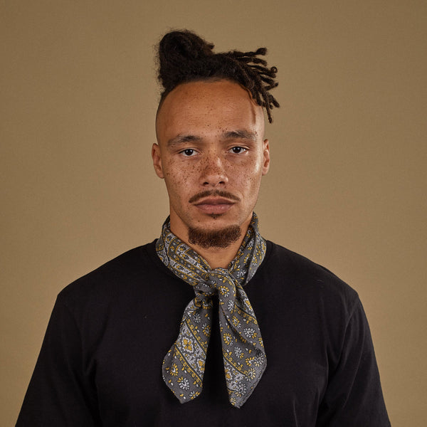 Top-half view of model looking straight ahead wearing 'The Whitehorse' grey and yellow multi-patterned bandana. Tied snugly around neck in a simple knot and paired with a black t-shirt.