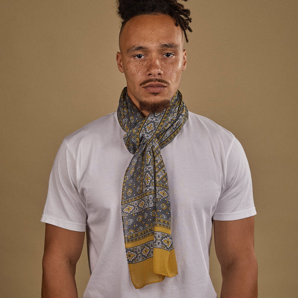 Close-up of model looking straight ahead wearing 'The Whitehorse' yellow, grey and white patterned wide bohemian scarf. Wrapped snugly around neck in a simple knot and paired with a white t-shirt.